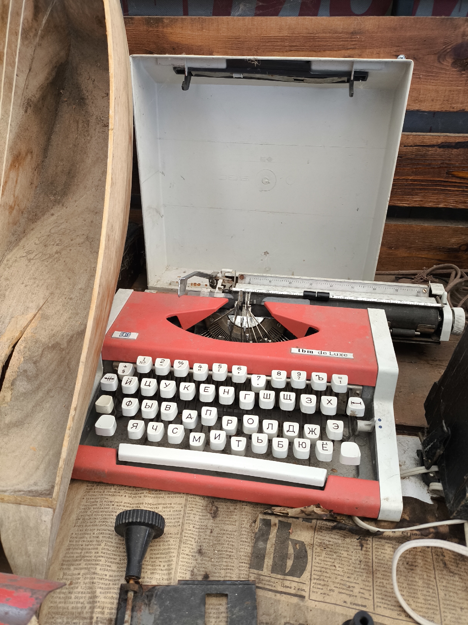 The image shows an old, red and white typewriter, with a white lid open, resting on a surface covered with newspapers. The typewriter is positioned at an angle, with the keyboard facing the viewer. The keys are white with black lettering, and some of them are partially obscured. To the left of the typewriter is a rustic wooden trough-like object, which appears to be made of light-colored wood and is partially filled with dirt and debris. The background consists of wooden planks and a dark object on the right side of the frame. The lighting is natural and appears to be from an overhead source. The scene is set in an outdoor or semi-outdoor environment, possibly a workshop or a storage area. The newspapers underneath the typewriter are aged and have visible text in a Cyrillic alphabet. The overall impression is one of age and disuse. No people are visible in the image.