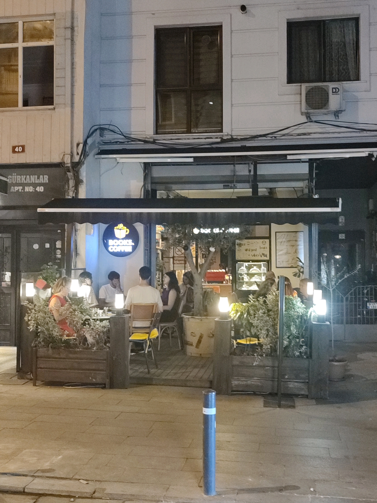 The image shows an outdoor seating area of a cafe in Kadıköy, Turkey, during the evening. The cafe is named "BOOKS, COFFEE," as indicated by a circular sign with a coffee cup graphic. Several people are seated at tables, some are eating or drinking, and others are engaged in conversation. The seating area is decorated with plants and lit by lamps. The facade of the building features windows and an air conditioning unit. A street sign indicating "40" and "GÜRKANLAR APT. NO: 40" is visible. The pavement is paved with stone tiles.