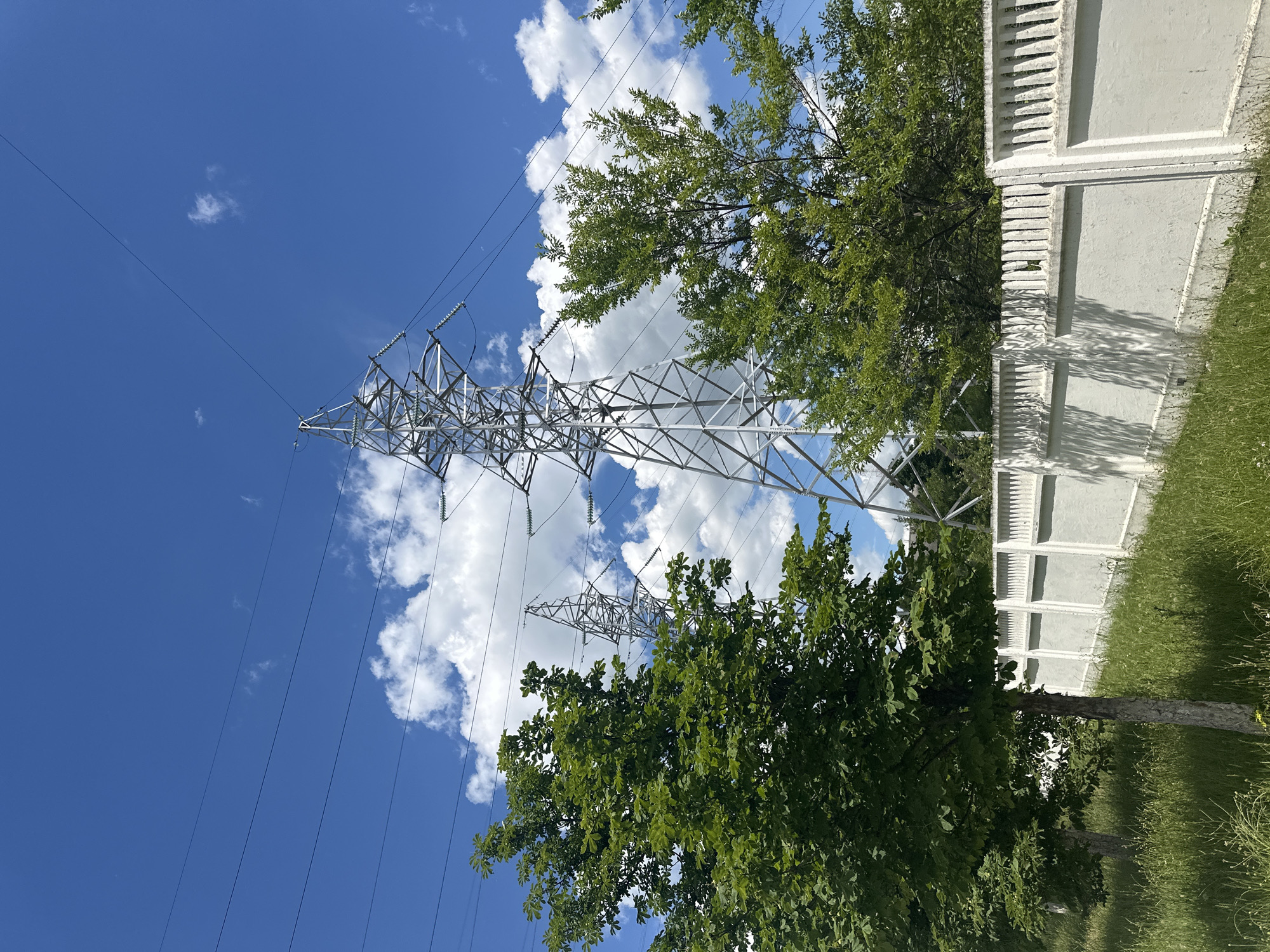 This is a photograph of a landscape with power lines and pylons against a bright blue sky with scattered white clouds. In the foreground, a white concrete fence runs horizontally across the image. To the left of the fence, there is tall green grass and two trees with lush green foliage. Behind the fence and trees, a large metal pylon is visible, supporting numerous power lines that stretch across the sky. Another similar pylon is partially visible further in the background to the left. The scene appears to be an outdoor area, possibly on the outskirts of a city, given the presence of industrial infrastructure like power lines. The lighting suggests it is daytime, likely mid-morning or afternoon. There are no people or animals present in the image. The overall impression is one of a sunny day with a juxtaposition of natural elements (trees, grass, sky) and man-made structures.