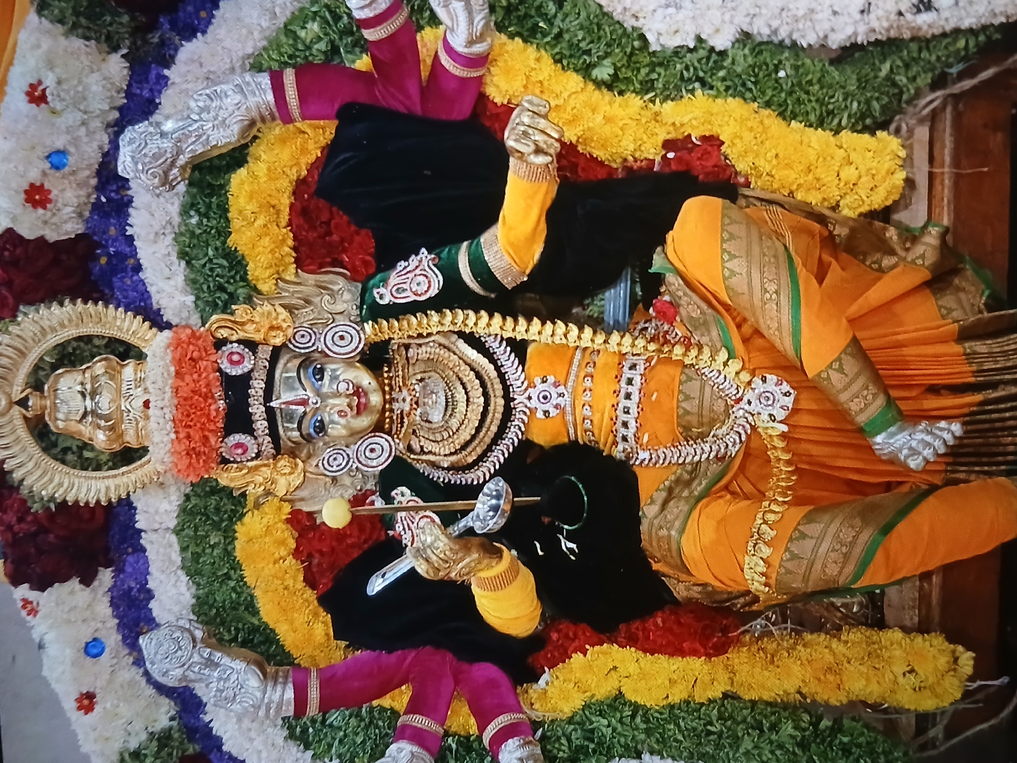 The image depicts a religious deity lying on a bed of flowers. The deity is adorned in gold jewelry and orange garments with green trim. The body is draped in black material. The feet are silver. A silver spoon is held in the deity's left hand. The background is a colorful arrangement of flowers. The image suggests a location in Bengaluru, India, given the South Indian style of the deity's adornment.