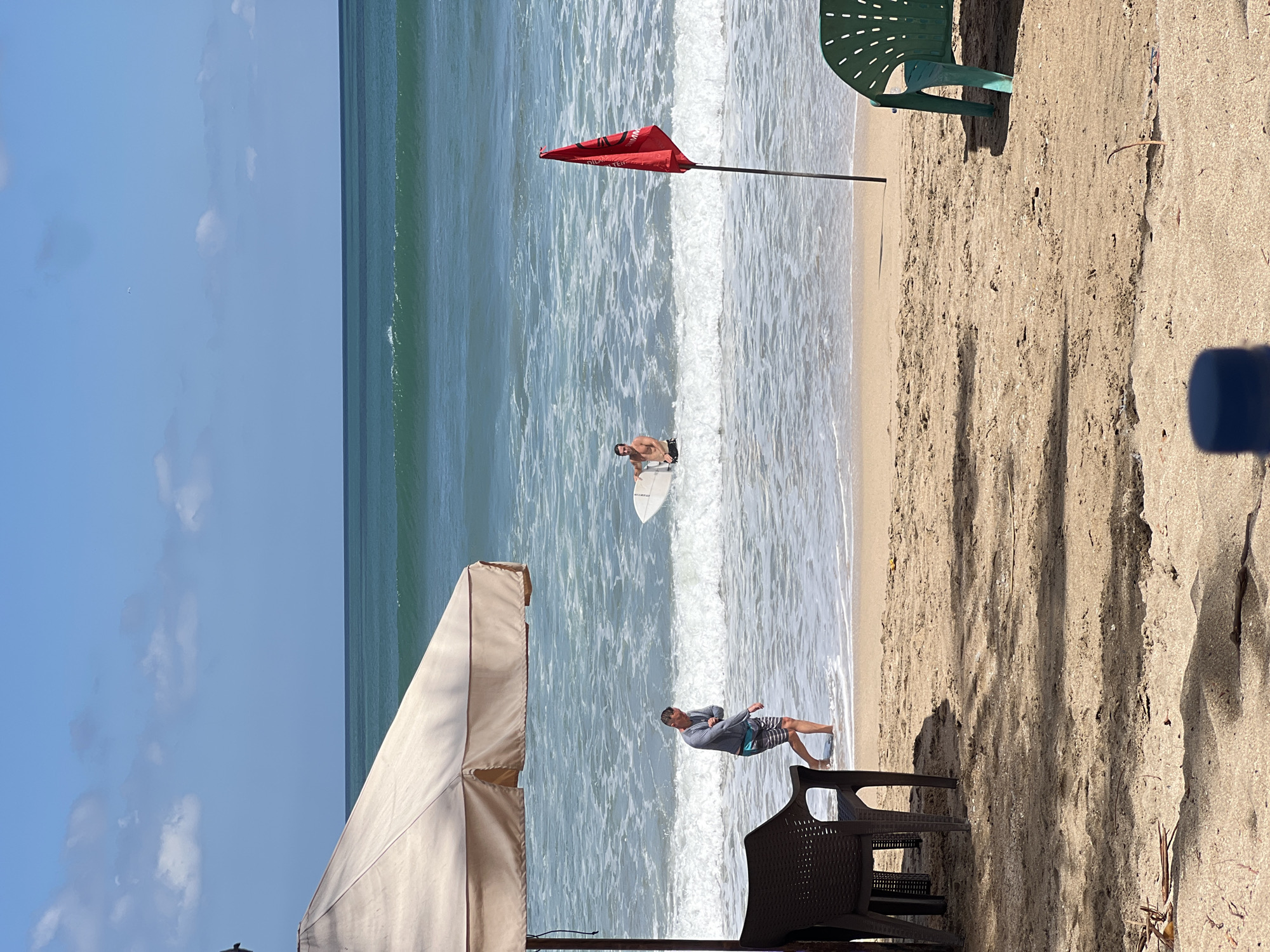 A scenic view of Kuta Beach in Kuta, Indonesia shows a sandy shore with two men in the water. The man on the left, wearing a grey long-sleeved shirt and patterned shorts, is wading in the shallow water. The man on the right, shirtless and wearing dark shorts, stands in the water with a surfboard. A red flag, signaling caution, is planted in the sand near the water's edge. In the foreground, a large beige umbrella provides shade over a dark plastic chair. To the right, a green plastic chair is partially visible. The ocean stretches out to the horizon under a clear blue sky dotted with white clouds.