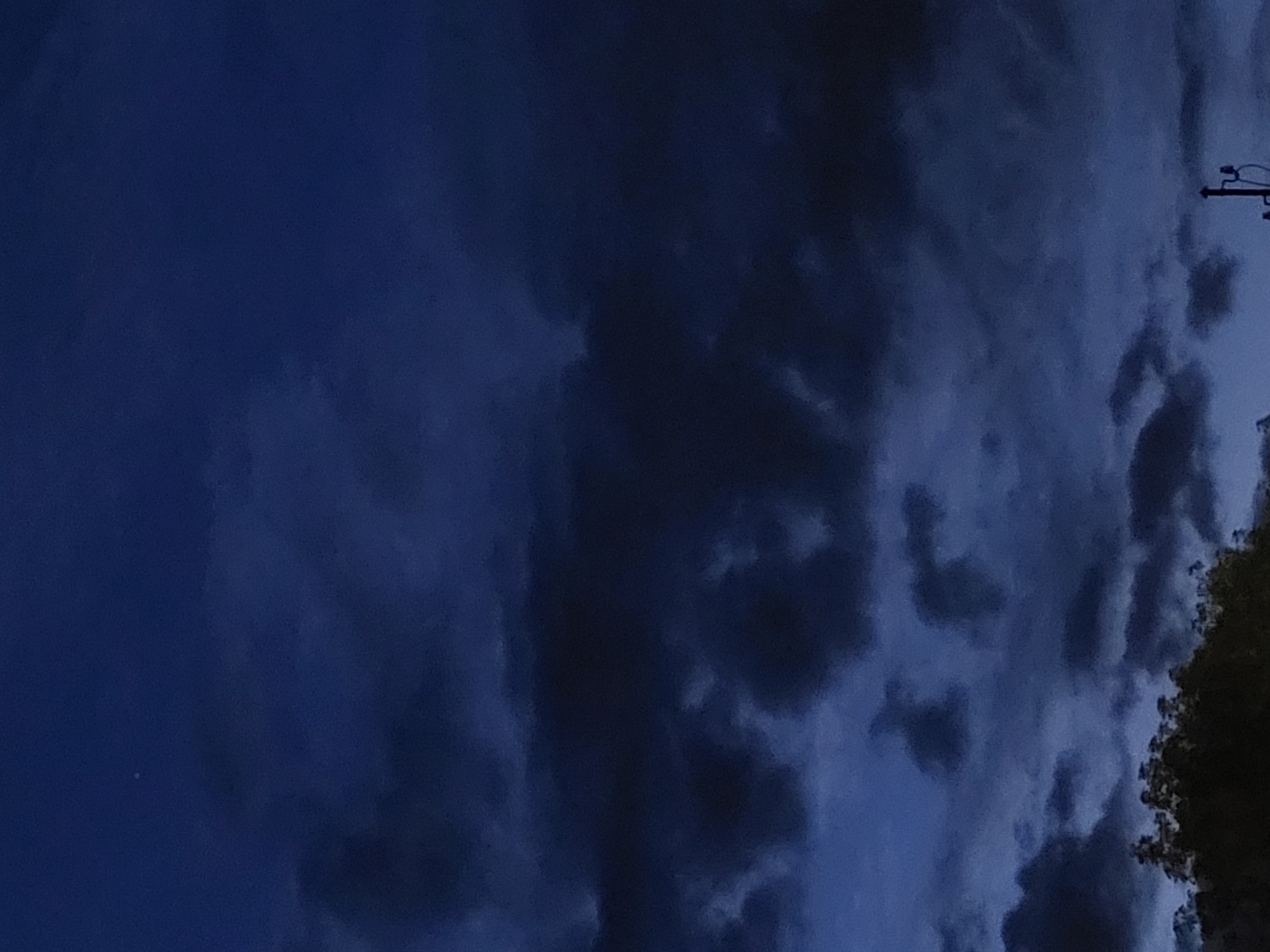 The image captures a twilight sky over a section of the city of Jelgava, Latvia. The sky is a deep, moody blue, filled with dark, textured clouds. In the upper portion of the image, a single, faint star is visible. Towards the bottom, the silhouettes of trees form a dark, dense line, suggesting a park or wooded area. A small portion of a utility pole or antenna structure is visible on the right side of the frame, piercing the lower edge of the cloud cover. The overall scene is quiet and atmospheric, indicative of the transition from day to night.