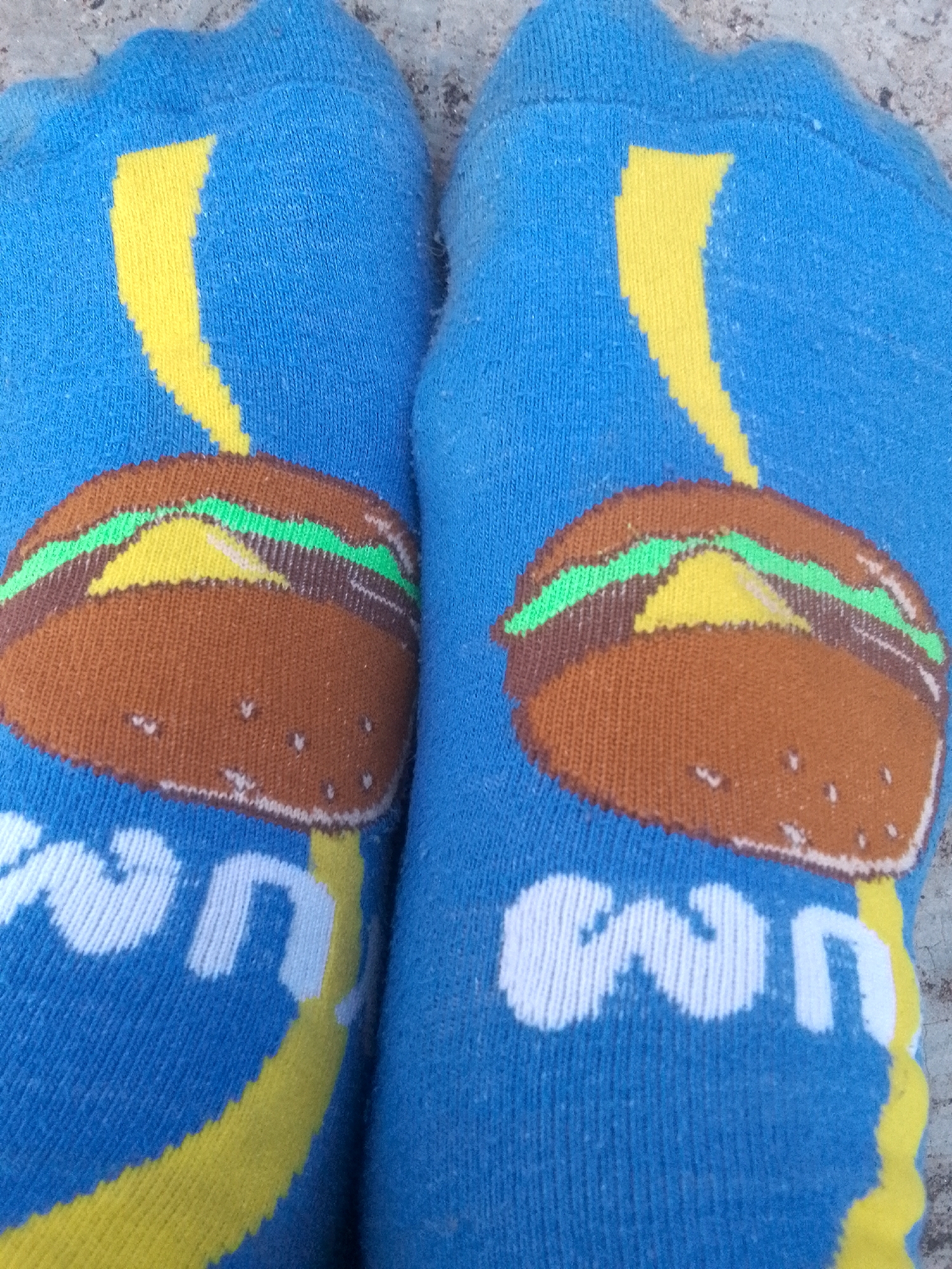 Two blue socks with a burger design on each. The burgers have brown buns, green lettuce, and yellow cheese. Underneath the burgers are the letters "WN" in white. A yellow streak runs vertically behind the burger on each sock. The socks are placed on a textured grey surface. Location: Altagracia, Venezuela is irrelevant to the image and is not depicted.
