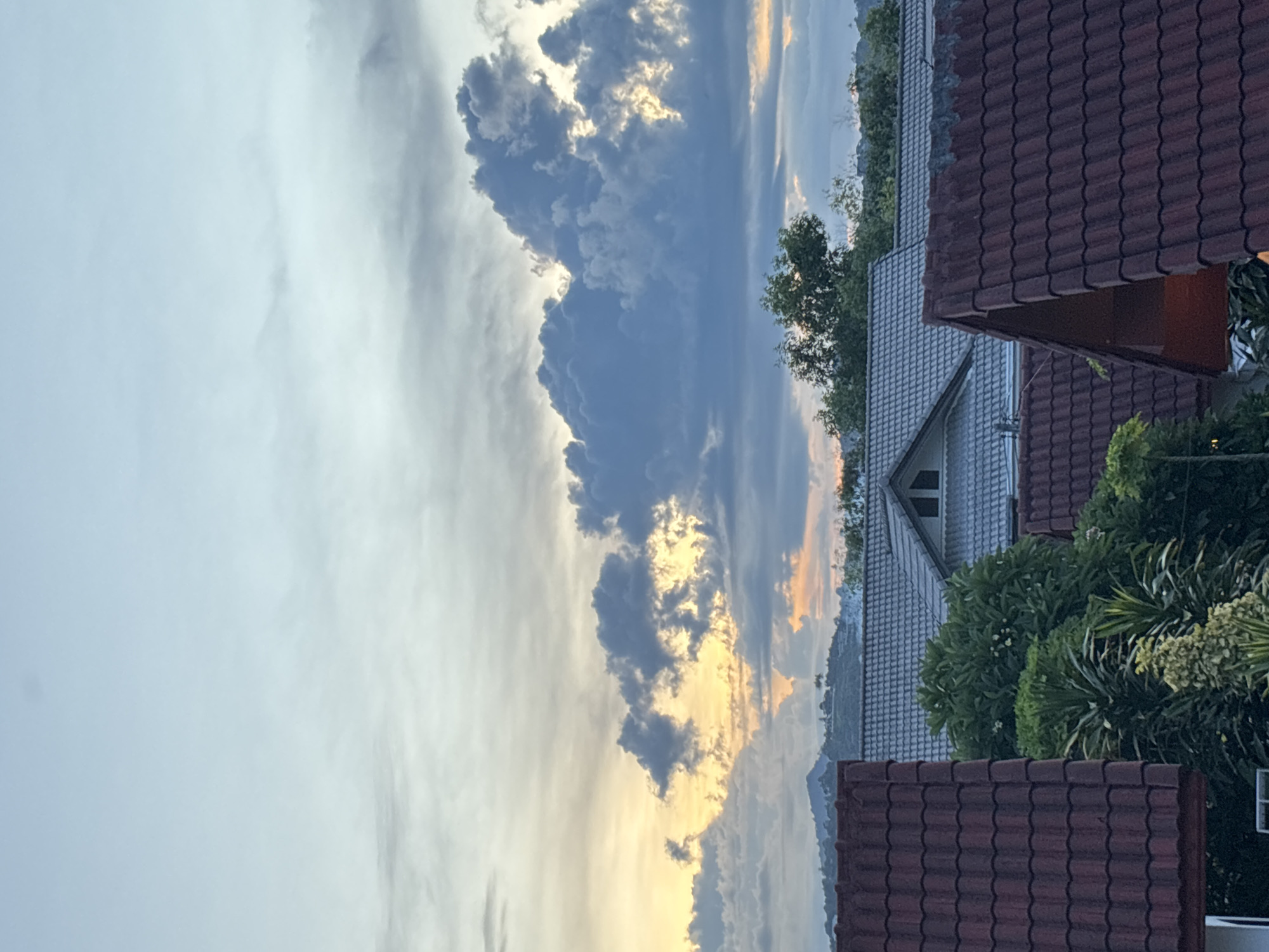 The image depicts a residential area at dusk, with a view of rooftops and a cloudy sky. In the foreground, several red-tiled roofs are visible. Behind them, lighter gray-tiled roofs of other houses are seen, one with a prominent triangular gable and a small attic window. Lush green trees and foliage are interspersed between the houses. In the background, a distant mountain range is silhouetted against the horizon. The sky is filled with dramatic clouds, with pockets of orange and yellow light from the setting sun peeking through, indicating either sunrise or sunset. The overall impression is one of a quiet, suburban evening.