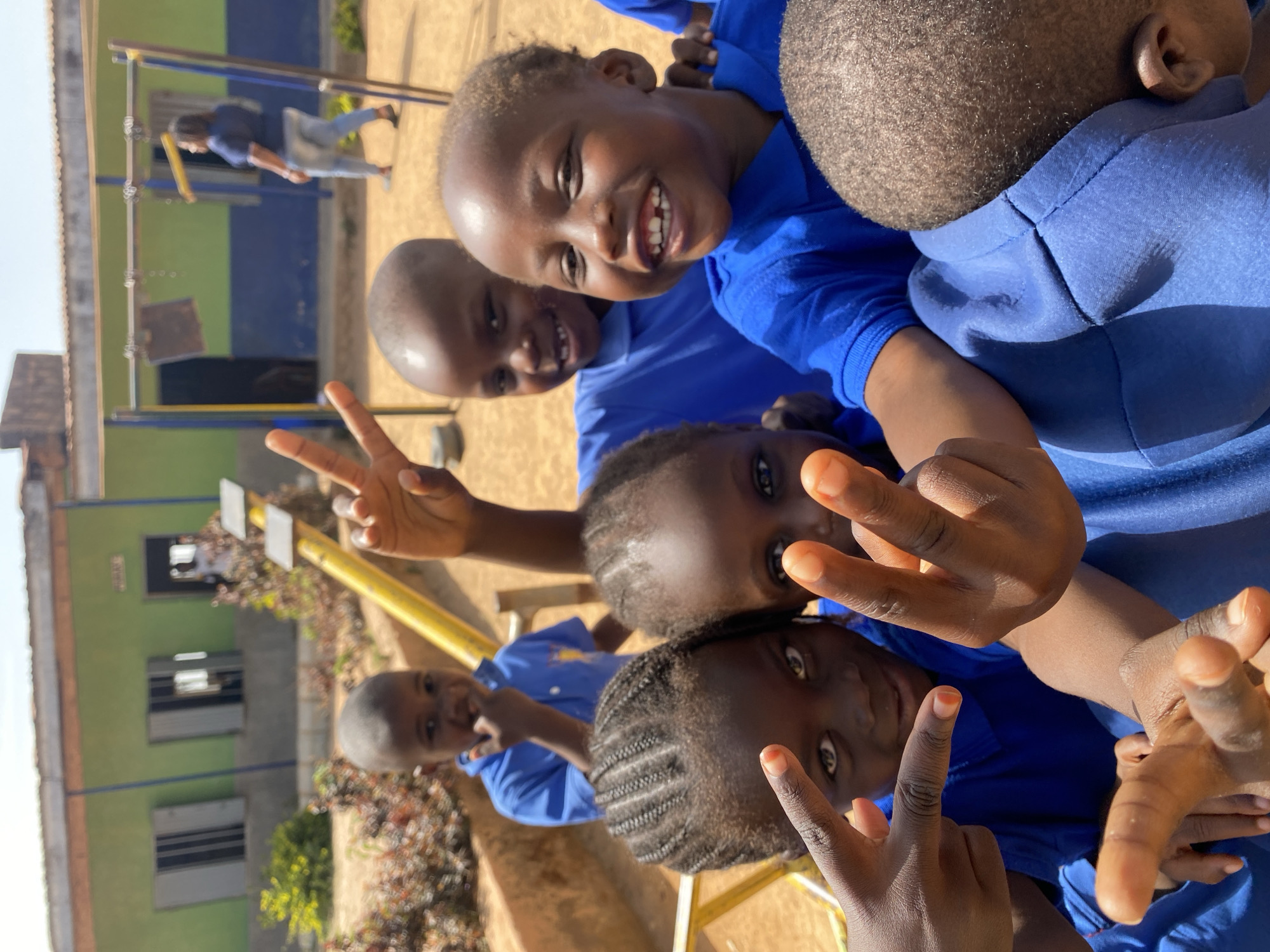 A group of young children wearing blue shirts are posing for a photo outdoors, near a school or playground in Gura Top, Nigeria. Some of them are making peace signs with their hands. In the background, there is a building painted in blue and green, as well as playground equipment. A person appears to be on the playground equipment. The scene is captured in broad daylight.