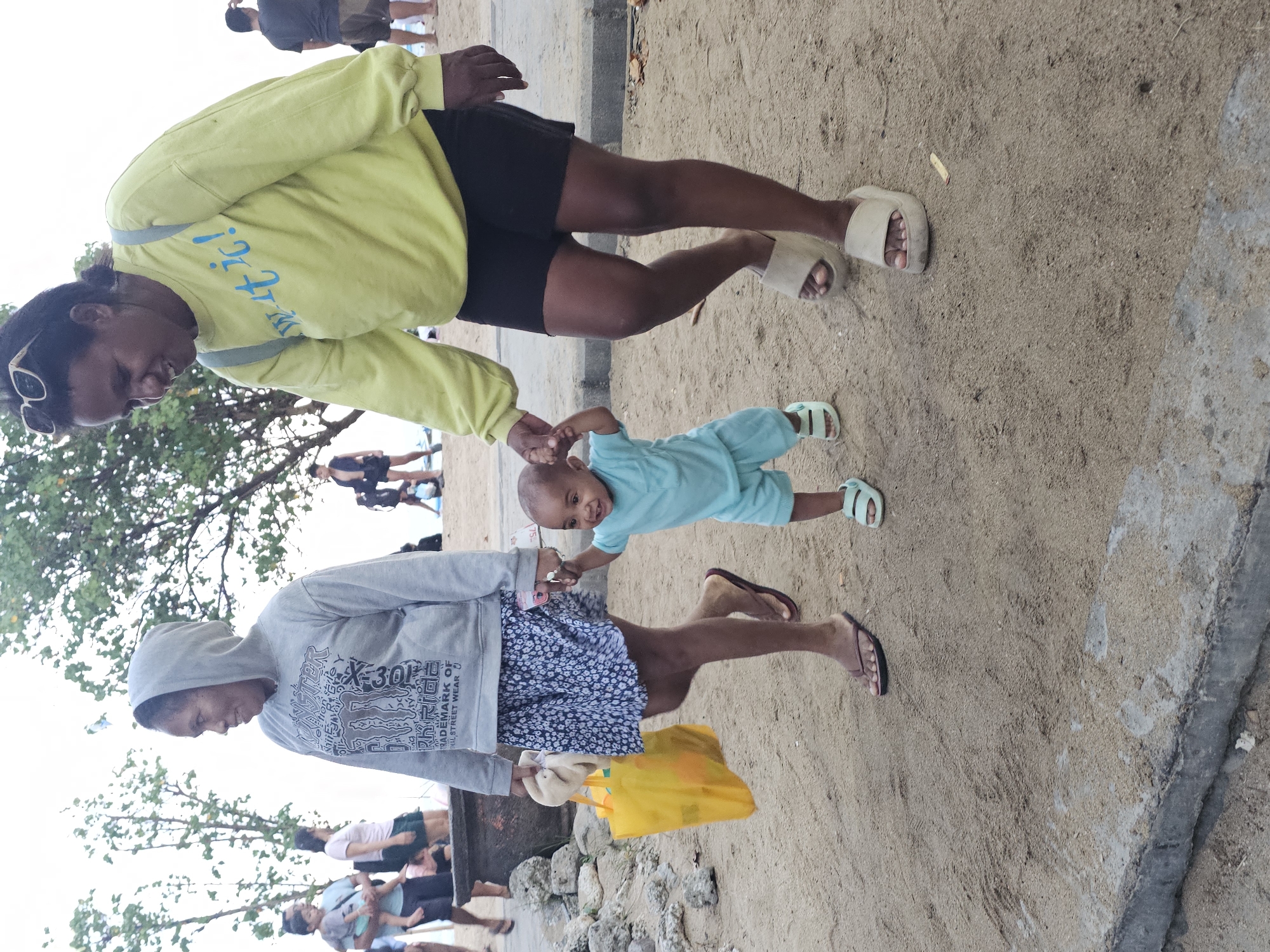 The image shows two adults and a child on a sandy area, likely a beach. One adult, wearing a yellow long-sleeved shirt, black shorts, and white sandals, holds the hand of a child dressed in blue. The child appears to be walking or being supported. The second adult, wearing a gray hooded sweatshirt and a blue and white patterned skirt, carries a yellow bag. In the background, other people are visible, suggesting a public space. The location is likely on a beach near Denpasar, Indonesia. The scene appears to be a casual outing.