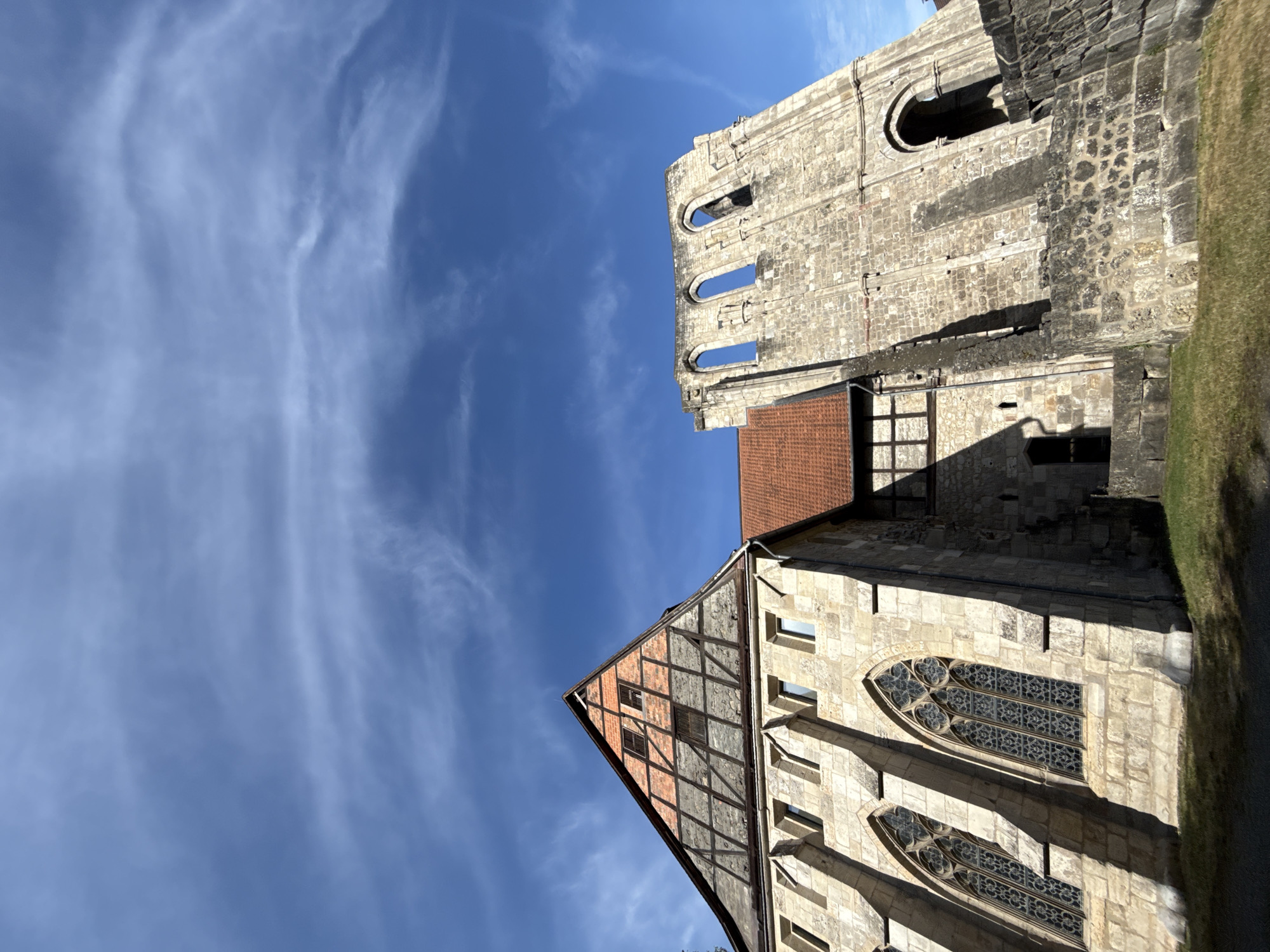 This is a low-angle, full-shot photograph of the exteriors of the Walkenried Abbey in Walkenried, Germany. The sky is a deep blue with wispy white clouds. On the left side of the frame, a section of the abbey building with a large, pitched roof covered in orange tiles is visible. This part of the building is constructed from large, beige stone blocks and features arched windows with intricate leaded glass designs. The roof section is a complex structure with exposed wooden beams and red brickwork visible on the gabled end. To the right of this section, a tall, stone ruin stands. This ruin is also constructed from rough-hewn stone blocks and has several large, arched openings without glass, suggesting it is a preserved remnant of a larger structure. The ground in front of the abbey is covered in green grass, with shadows cast by the buildings. The architecture indicates a historical religious site, likely a monastery or church. There are no people visible in the frame.