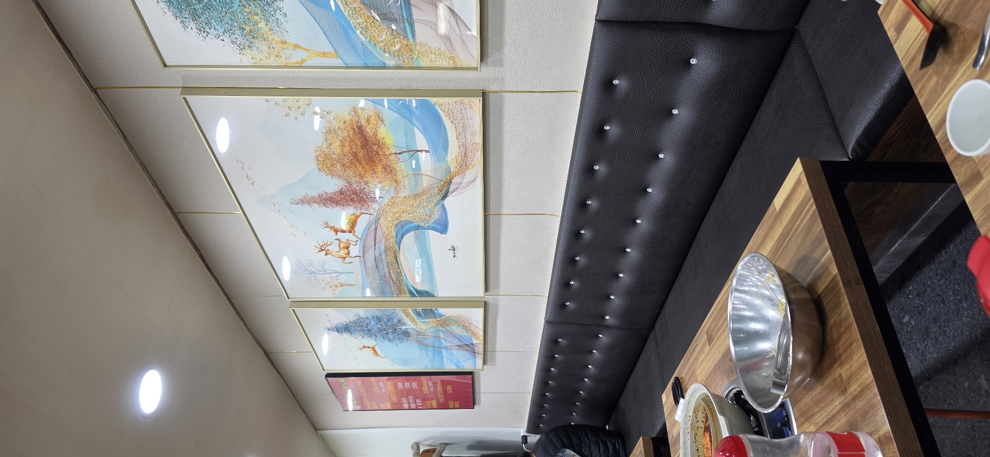 The image shows the interior of a restaurant in Cheongju-si, South Korea. The scene is dominated by a long, dark leather bench with tufted diamond patterns, typical of booth seating in Korean restaurants. In front of the bench, a wooden table is visible, with metal legs. On the table, there are several items, including a metal bowl containing some food and utensils, a clear plastic bottle with a red lid, and another smaller metal bowl with tongs inside. In the background, a wall is adorned with three large, framed abstract paintings. The paintings feature stylized landscapes with deer, trees, and flowing abstract elements in shades of blue, gold, and orange. To the left of the paintings, a smaller red sign with Korean text is mounted on the wall. The ceiling has recessed lighting. The overall impression is that of a casual dining establishment.