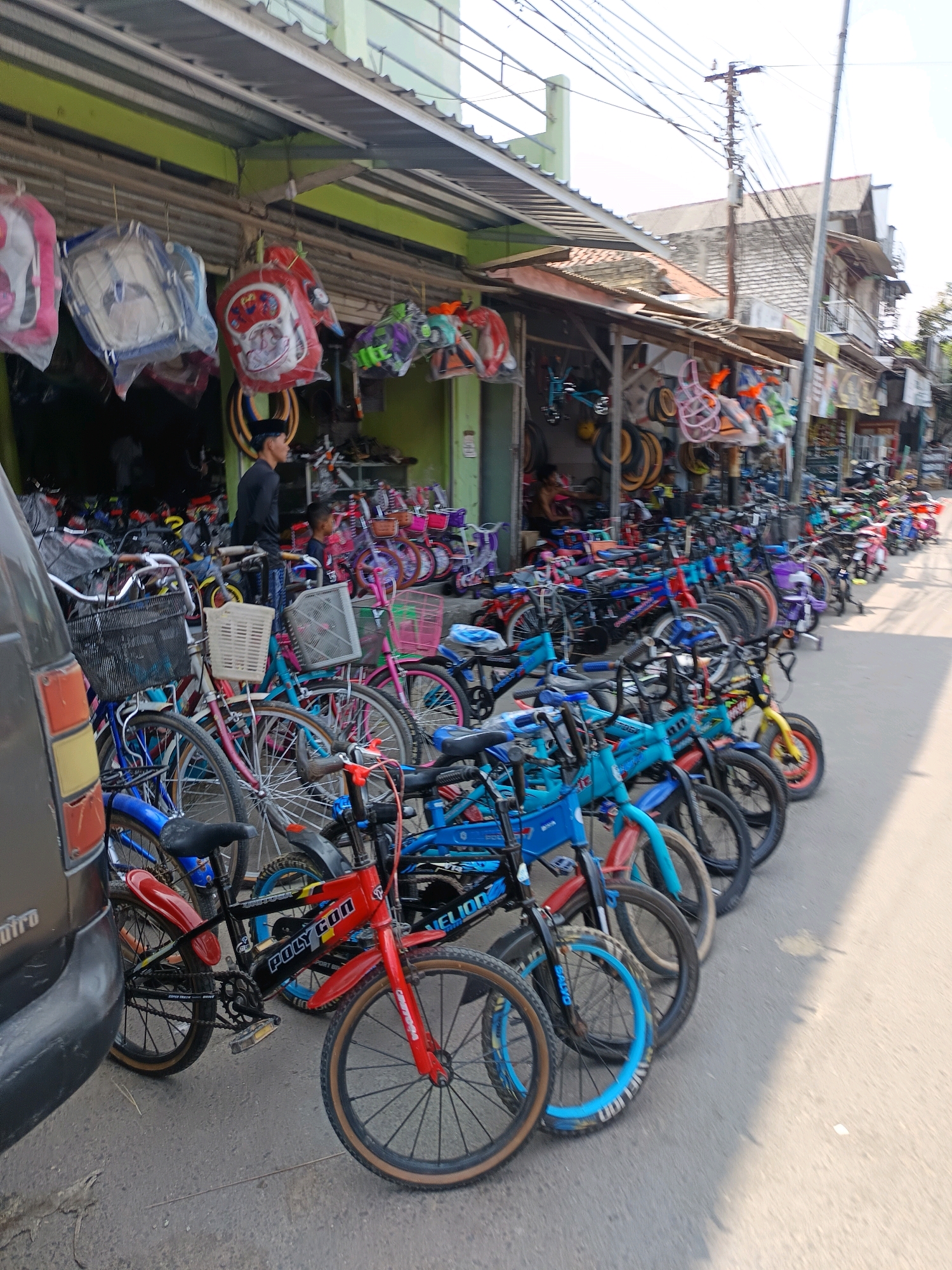 A street-level view of a bicycle shop in Blega, Indonesia, is depicted. The shop is lined with various bicycles of different sizes and colors, parked on the sidewalk and extending into the street. The facade of the shop is covered with a green awning. Several backpacks are hanging from the awning, some in plastic packaging. A man in a black shirt and head covering is standing in the doorway of the shop, looking to the right. The street is paved and appears to be busy with activity. The lighting suggests it is daytime, with sunlight casting shadows. The overall scene conveys a typical commercial setting in a Southeast Asian town.