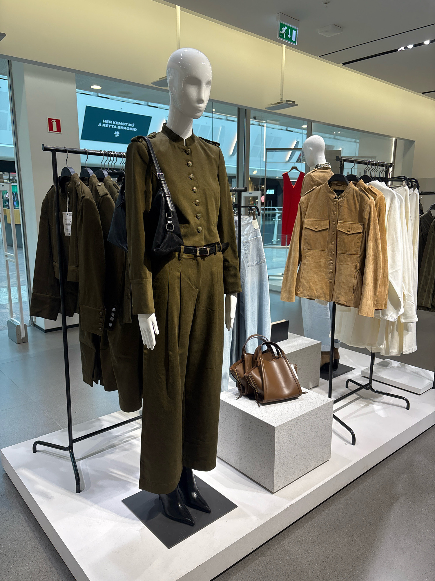 This image depicts a retail store display featuring mannequins and clothing racks in Kopavogur, Iceland. The primary focus is on a mannequin dressed in a khaki green, military-inspired outfit, complete with a matching belt and black heeled boots. To the right, another mannequin is partially visible, wearing a tan suede jacket. Several clothing racks are laden with garments, including more khaki green jackets, light blue jeans, a red dress, tan suede jackets, and white tops. Two brown handbags are displayed on pedestals in the foreground. An exit sign is mounted on the ceiling, and a fire extinguisher sign is visible on the wall in the background. The store appears modern, with large windows and a clean, minimalist aesthetic. The overall scene is one of a fashion retail environment.