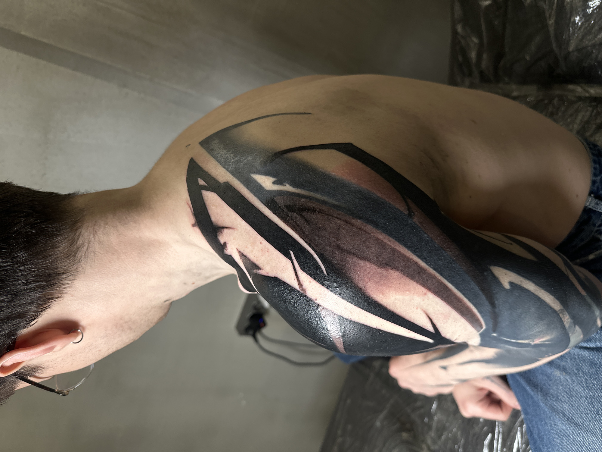 The image shows a man's left arm with a large, primarily black abstract tattoo that covers the shoulder and extends down the arm. The tattoo incorporates black, grey, and some skin-toned areas within the design. The man is wearing blue jeans. He has short dark hair, glasses, and an earring. The background includes a grey wall and a surface covered with a protective plastic sheet. The location is not discernible, so it is not possible to identify if it is in Moscow, Russia.
