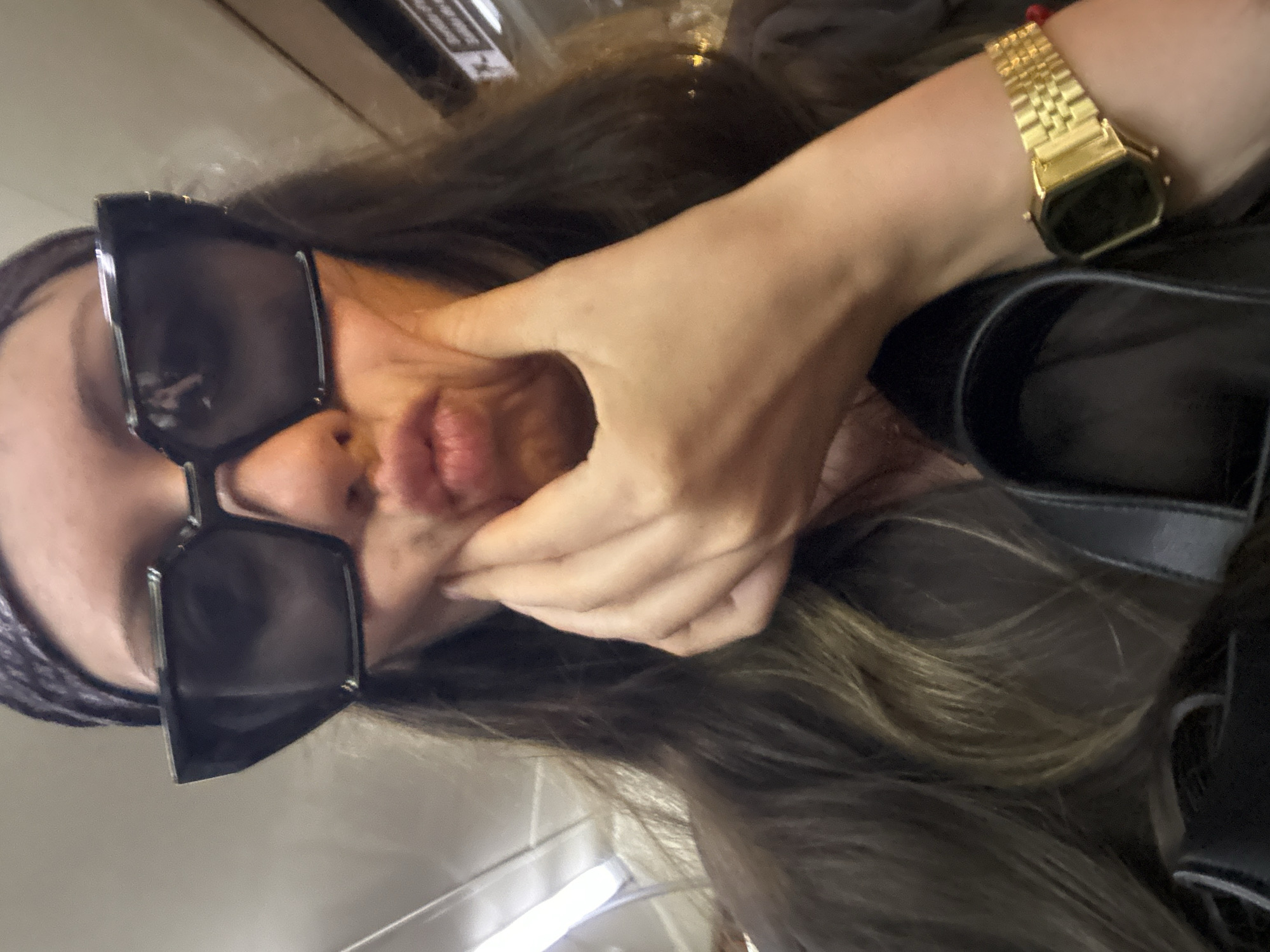 A young woman with long brown hair and a black bandana covering her head, wearing large, angular, dark sunglasses, makes a duck face while holding her cheeks with her left hand. She is wearing a gold-colored digital watch on her right wrist. She is wearing a dark colored jacket. The background is blurred and appears to be a public indoor space, possibly a train or subway car, given the presence of fluorescent lighting and signage. The overall lighting is dim. There are no discernible landmarks or context clues that suggest the location is Barcelona, Spain.