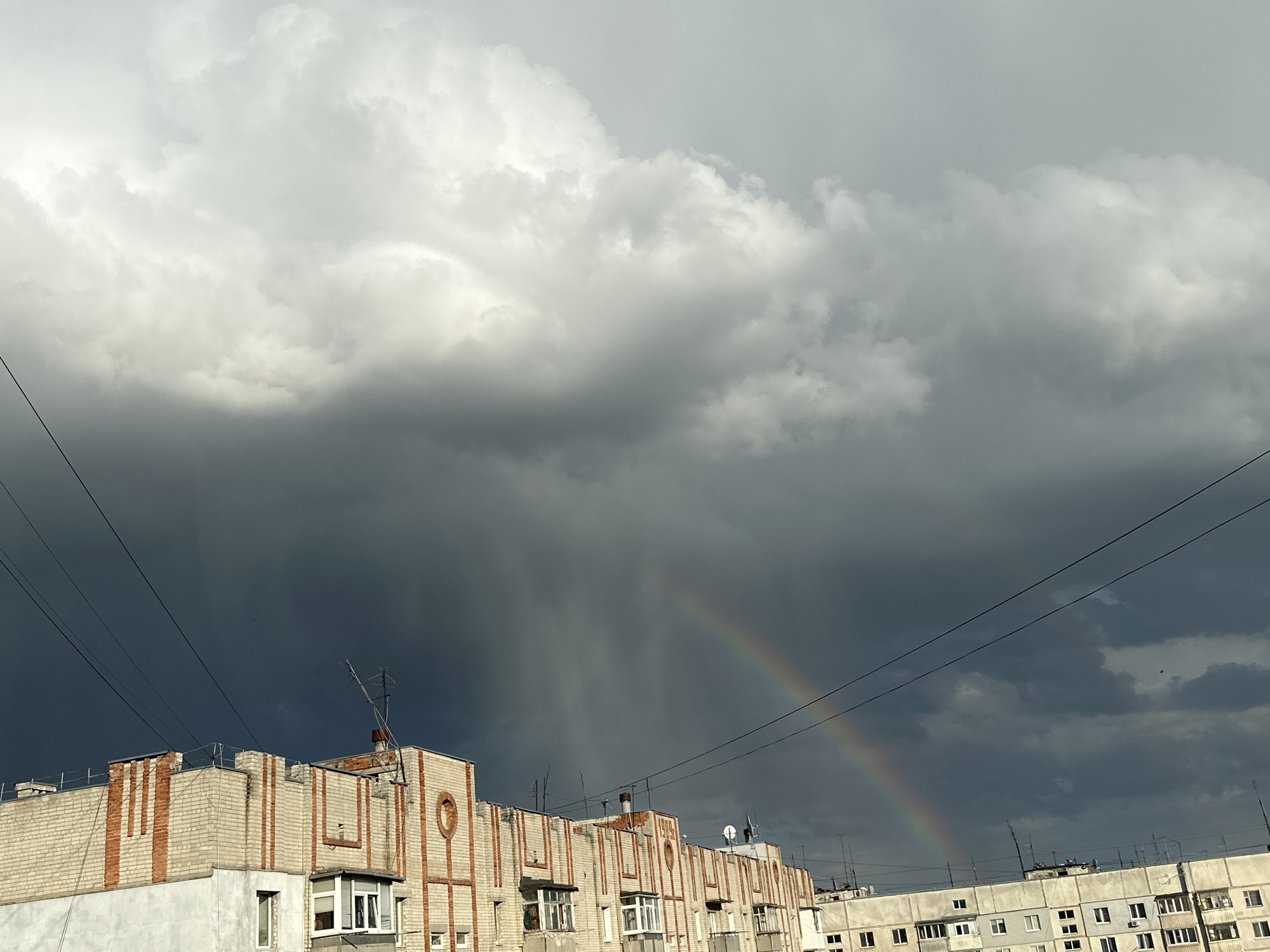In the city of Unknown, Unknown, a dramatic sky unfolds above a row of apartment buildings. Dark, heavy storm clouds dominate the upper portion of the frame, with lighter, fluffy clouds scattered towards the top. A faint rainbow arcs across the sky, partially obscured by the dark clouds, hinting at recent rainfall. The apartment buildings are constructed of light-colored brick with decorative reddish-brown stripes. Some buildings have visible antennas and satellite dishes. Utility wires stretch diagonally across the scene. The overall impression is one of a fleeting moment of natural beauty amidst urban architecture, under a dynamic and changing sky.