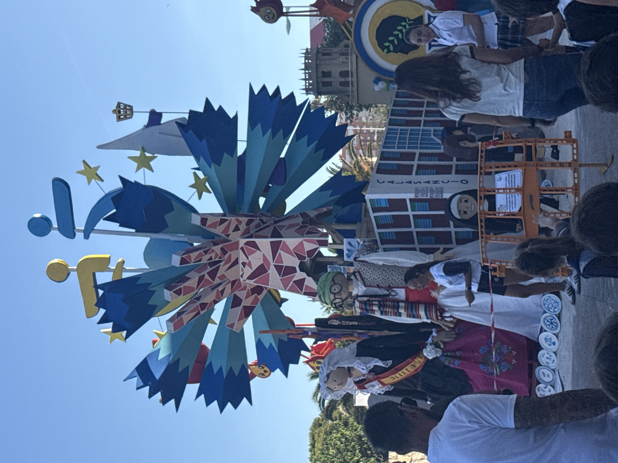 The image depicts a vibrant outdoor scene, possibly a festival or a public event, featuring large, colorful figures and elaborate decorations. In the foreground, a man in a white t-shirt is seen from the back, with tattoos visible on his arm. To his right, several children are partially visible, and a woman with long dark hair is walking away from the camera.
The central focus of the image is a towering, abstract sculpture made of geometric shapes and vibrant colors, predominantly blue, red, and yellow, with some mosaic-like detailing. Attached to this structure are various figures, including a large doll dressed as a nun and two other larger-than-life figures. One of these figures is a woman wearing a black dress with a white ruffled collar and a sash that reads "BILBAO." She is adorned with a sash and a veil. Another figure to her right is a man wearing a green hat and glasses, a patterned vest, and a striped scarf. In front of these figures, there is a white fence with orange safety barriers. A young woman in a white t-shirt and dark shorts is standing and gesturing, seemingly interacting with the scene. Another young woman in a white shirt and plaid skirt is visible in the background, smiling.
Behind the main display, there is a building with castle-like architecture. The sky is clear and blue, indicating daylight. Several palm trees and other greenery are visible, suggesting a warm climate. The ground appears to be paved. Decorative plates with blue patterns are arranged in a semi-circle in front of the large figures. The overall atmosphere appears festive and celebratory.