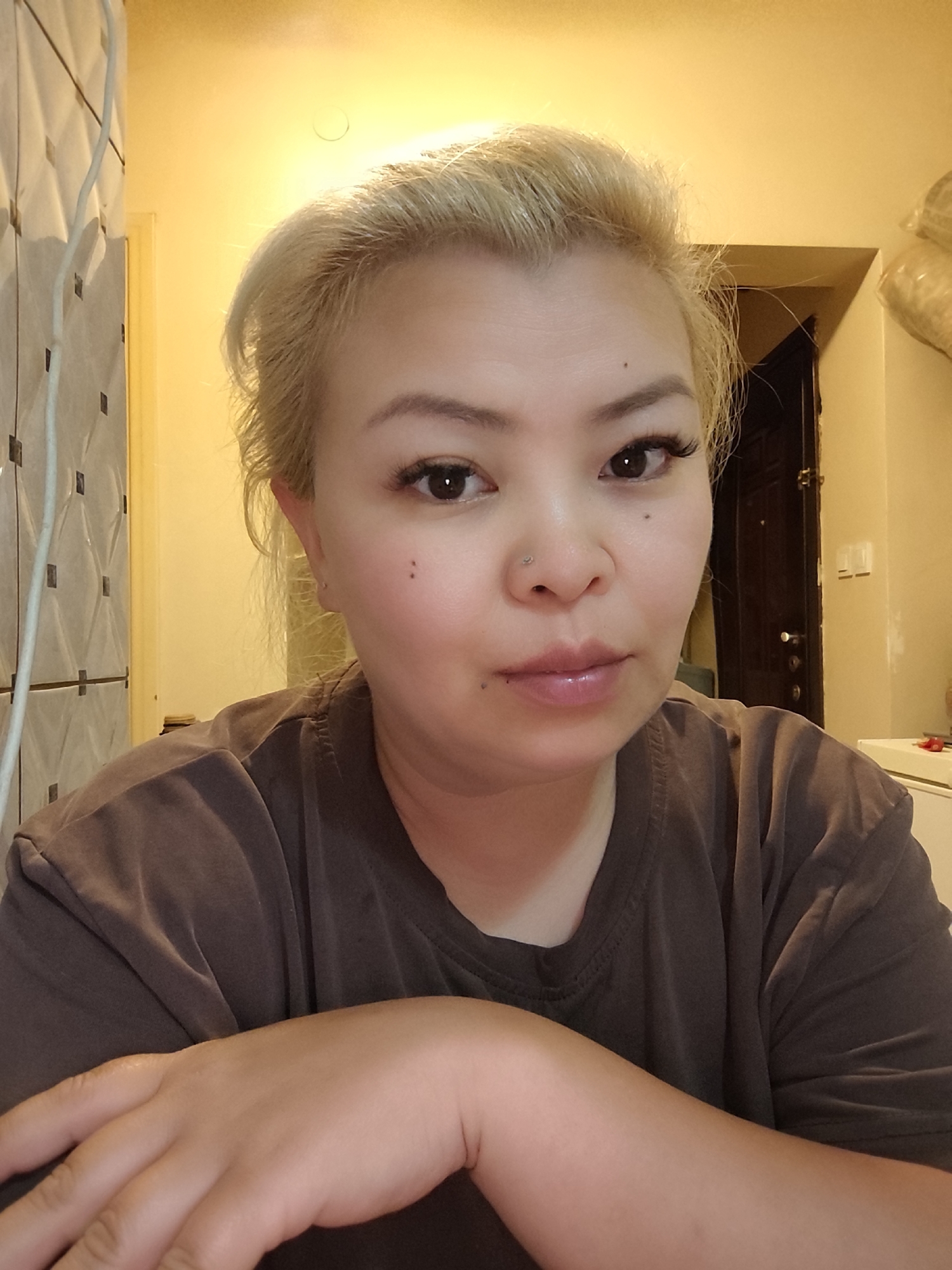 A close-up indoor portrait features a woman with light blonde or bleached hair, possibly of East Asian or Southeast Asian ethnicity. She has dark brown eyes, subtle makeup including eyeliner and what appear to be false eyelashes, and a small silver nose piercing on her left nostril. Two small dark spots are visible on her left cheek and one on her right, near her lower lip. She looks directly at the camera with a neutral to slightly pleasant expression, her lips slightly parted. She is wearing a dark brown or charcoal-colored t-shirt, and her arms are resting on a surface in front of her, with her right hand visible in the foreground.
The background is dimly lit with warm, yellowish artificial light, suggesting an indoor setting, likely in the evening or at night. To the left, a light-colored wall or partition with a grid of small dark squares is visible, alongside a white cable running vertically. To the right, a dark wooden door with a handle and lock is partially visible, leading to a darker interior space. Above the door, a plain light-colored wall can be seen, and in the upper right corner, some bundled, soft objects are stored. Below the door, a white surface, possibly a counter or piece of furniture, is partially visible. A circular light fixture cover is visible on the yellowish ceiling. The woman appears to be posing for a photograph, possibly a selfie, in what seems to be a casual home environment in Sarıyer, Turkey. There is no visible text in the image.