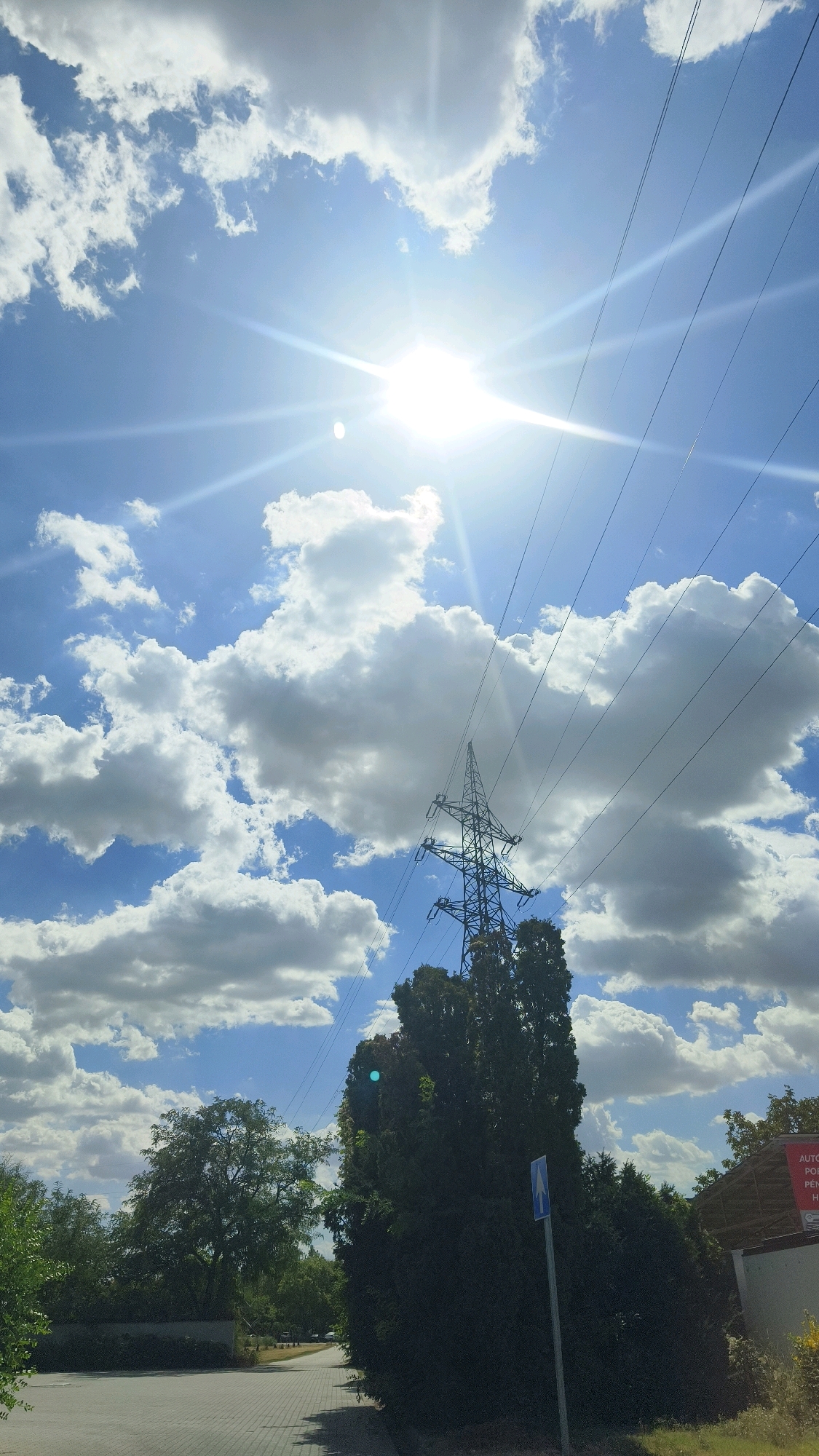 This is a photograph of a bright, sunny day with a clear blue sky filled with puffy white clouds. The sun is prominently featured in the upper center of the image, with strong lens flare effects. Power lines are visible crossing the sky, strung between transmission towers. In the mid-ground, a large lattice-style transmission tower stands tall amidst dense green trees. A paved walkway or road is in the foreground, with more trees and bushes lining the side. On the right side of the image, part of a building with a sign is visible, and a blue sign with a white arrow pointing upwards is planted in the ground near the trees. The scene appears to be an outdoor area in a city or suburban environment, possibly near a park or residential area. The specific location in Szeged, Hungary, cannot be determined from the visual information provided.