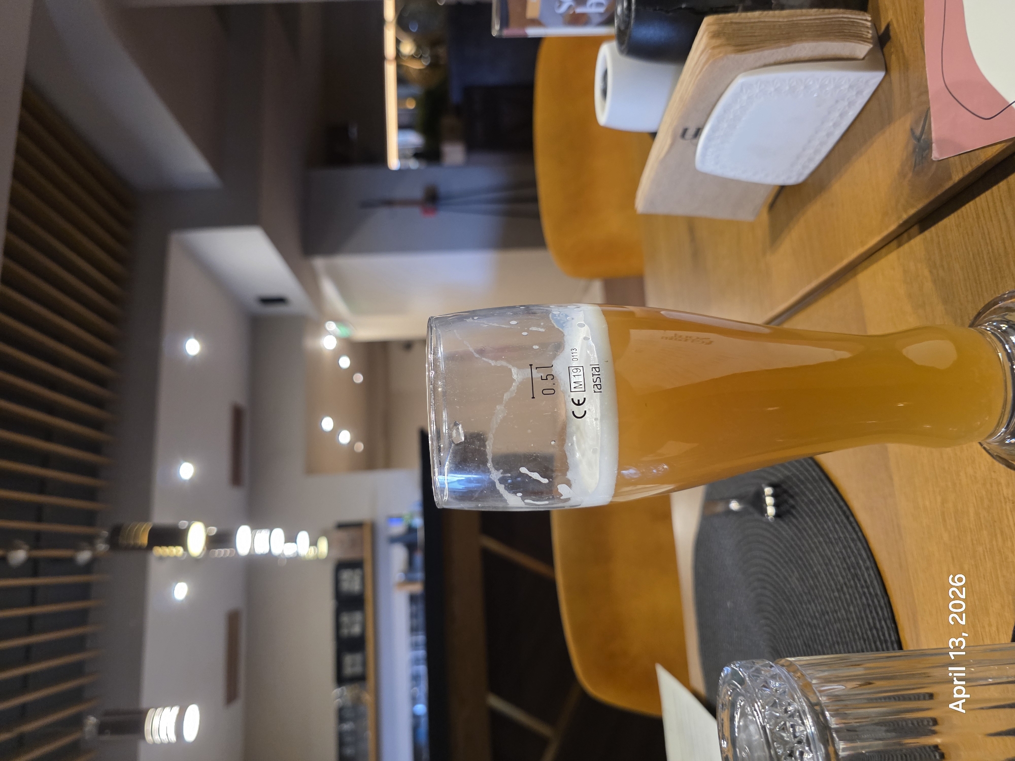 The image shows a tall beer glass, partially filled with a light-colored beer, sitting on a wooden table in what appears to be a restaurant or bar. There is a black circular placemat visible beneath part of the glass. Nearby on the table are a white coaster, a book, and other unidentified objects. In the background are orange chairs and a blurred view of the interior of the establishment, including lighting fixtures on a wall. The date "April 13, 2026" is watermarked on the bottom right. Without additional context, pinpointing the exact location in Sofia, Bulgaria, is not possible.