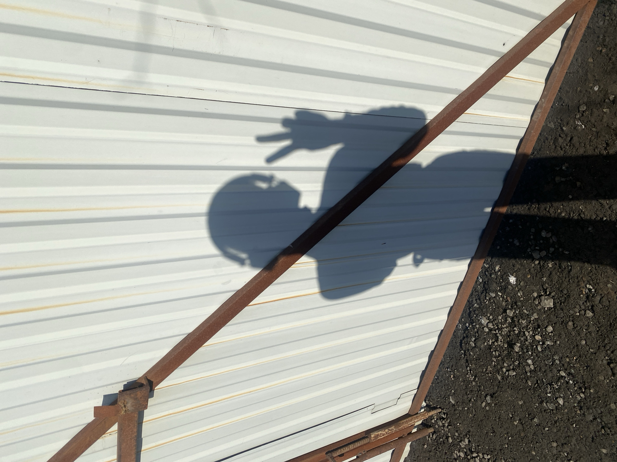 A high-angle shot depicts a weathered metal sheet wall, possibly part of a building, set in Unknown, Unknown. The wall, painted a light color with horizontal grooves, is marked with streaks of rust or discoloration. The shadow of a person is projected onto the wall, showing the individual's head and torso, with one hand raised, gesturing a "peace" or "victory" sign. A rusted metal frame structure is attached to the wall, with one corner resting on dark soil mixed with small stones. The scene is brightly lit, casting clear shadows.