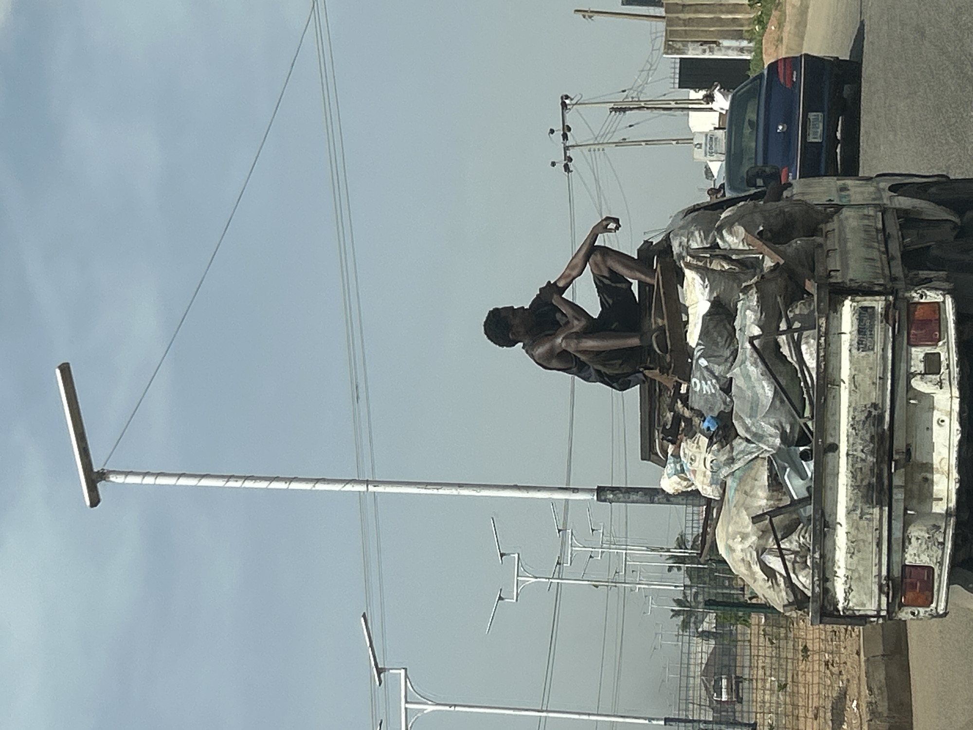 A man sits atop a pickup truck laden with bags of waste material on a road in Jos, Nigeria. The truck is white and visibly dirty. A dark blue car is behind the truck. The background includes streetlights and power lines against a hazy sky. Vegetation is visible in the distance, and a partially seen structure suggests an urban area.