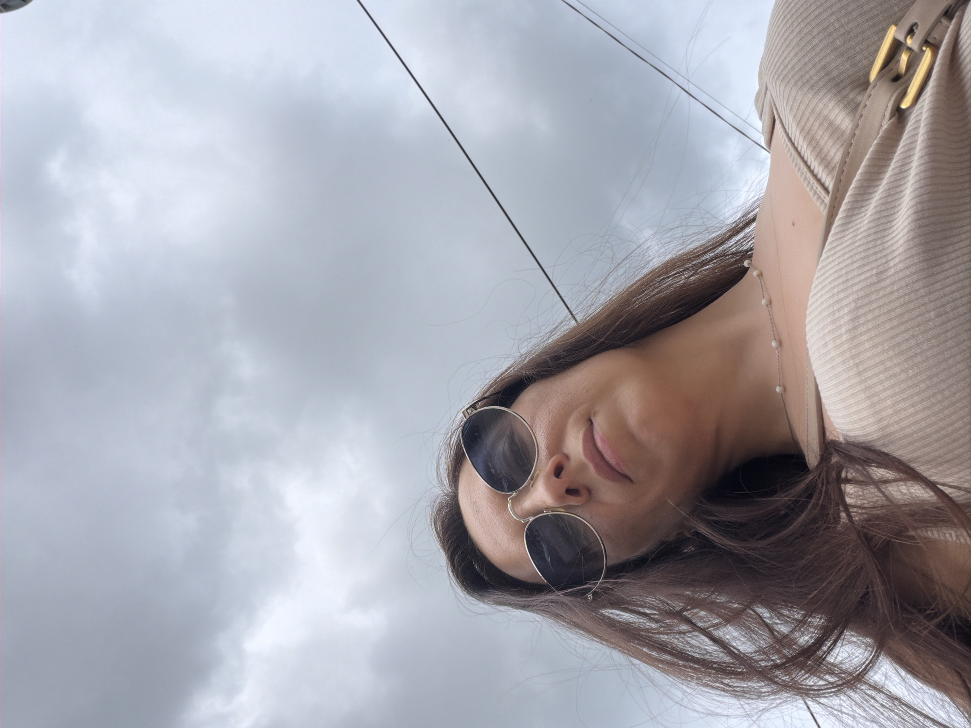 A low-angle, medium shot captures a woman with long, dark brown hair, wearing sunglasses and a light beige, ribbed top. She is looking down, and her face is partially obscured by her hair. The background is a cloudy sky with some telephone wires visible. The woman is positioned in the foreground, with her upper body filling most of the frame. The image appears to be taken outdoors, given the sky and overhead wires.