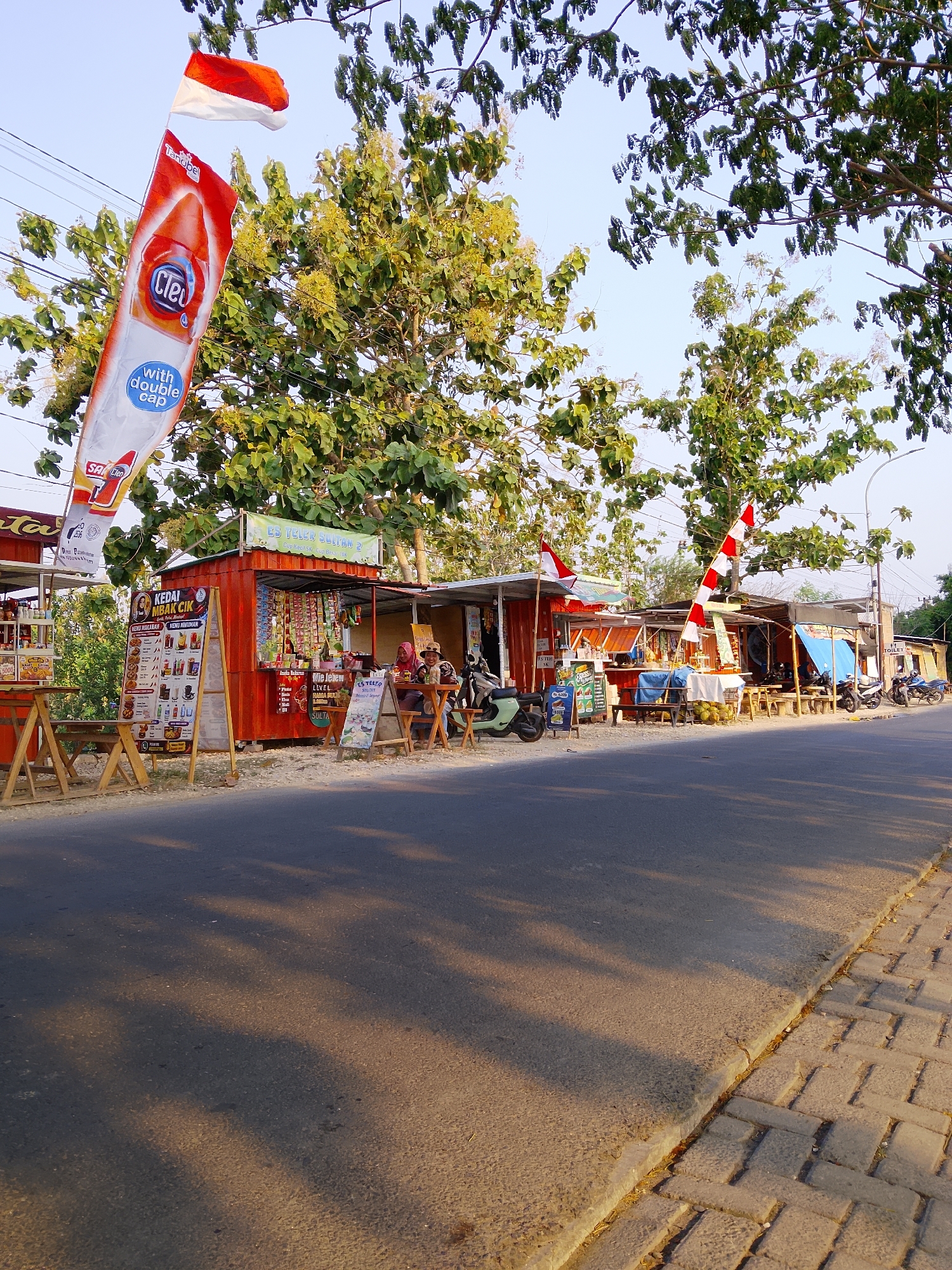 The image captures a street scene in Ngadrin, Indonesia, featuring a row of small businesses, likely food stalls or kiosks, set up along the roadside. The stalls are constructed from shipping containers and simple wooden structures, some adorned with Indonesian flags. In the foreground, a paved road runs alongside a sidewalk. Shadows from trees stretch across the road, indicating it is likely late afternoon.
One prominent stall displays a menu board for "KEDAI MBAK CIK" offering various food and drink options. Another stall, labeled "ES TEH SULTAN 2," appears to be selling beverages. A few individuals are visible, one sitting at a table outside a stall, seemingly interacting with another person. Several motorcycles are parked near the stalls, a common mode of transportation in the area.
A large banner advertising "Cleo" bottled water with a double cap is prominently displayed, attached to a pole. The overall atmosphere suggests a typical street scene in a town or village in Indonesia, with local commerce and daily life taking place outdoors. The backdrop consists of lush green trees and a clear sky.