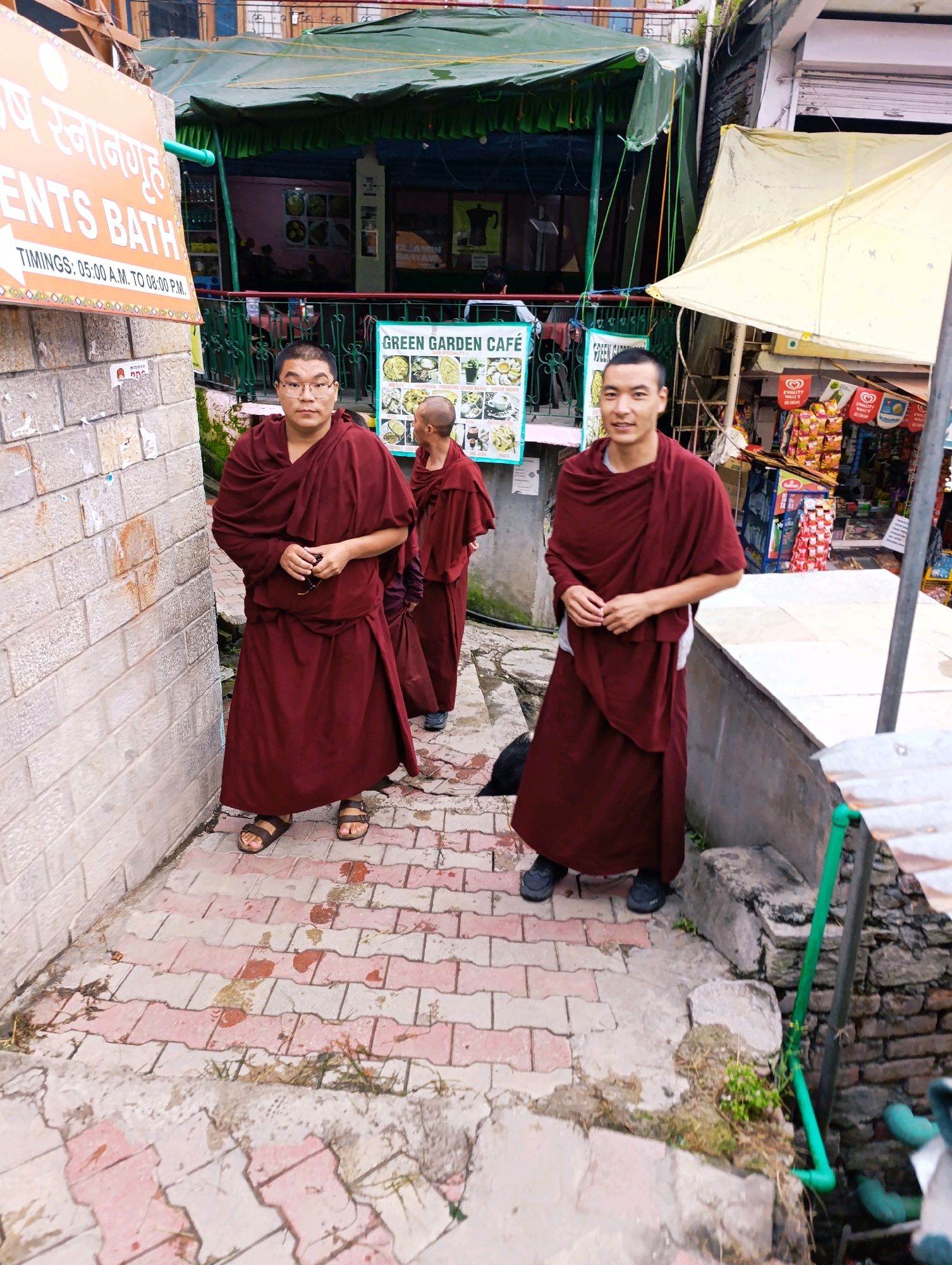 In Vashisht, India, three Buddhist monks in maroon robes stand on a paved walkway. The monk on the far left, wearing glasses, faces the camera. To his right, another monk, partially obscured, looks towards the right. The monk on the right, with short hair, smiles at the camera. Behind them, a green awning shades a building with a sign that reads "GREEN GARDEN CAFE". Further to the right, a market stall displays various packaged goods. The walkway is made of interlocking red and grey bricks, with some scattered debris and plants. A brick wall is visible on the left, and a stone structure with a green pipe is on the right.