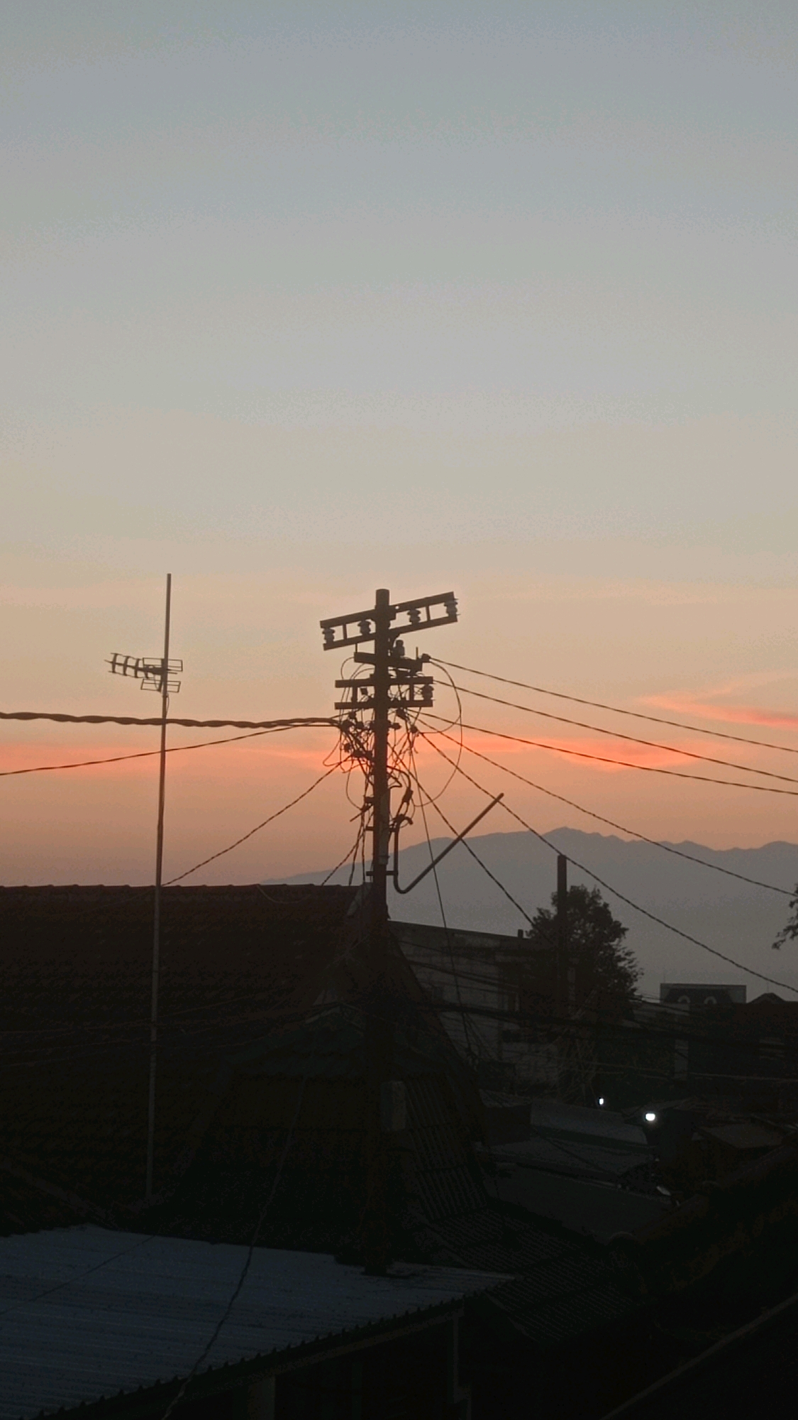 A silhouetted telephone pole with numerous tangled wires stands prominently in the foreground against a dusky sky. To the left, another pole with an antenna is visible. The sky transitions from a pale blue at the top to a soft orange hue near the horizon, suggesting either sunrise or sunset. In the mid-ground, a dark mass of rooftops indicates an urban or semi-urban environment. Distant, hazy mountains form the backdrop. The overall scene captures a moment in the city of Mulyoagung, Indonesia, characterized by its utility infrastructure and atmospheric lighting. There are no visible people in the image.
