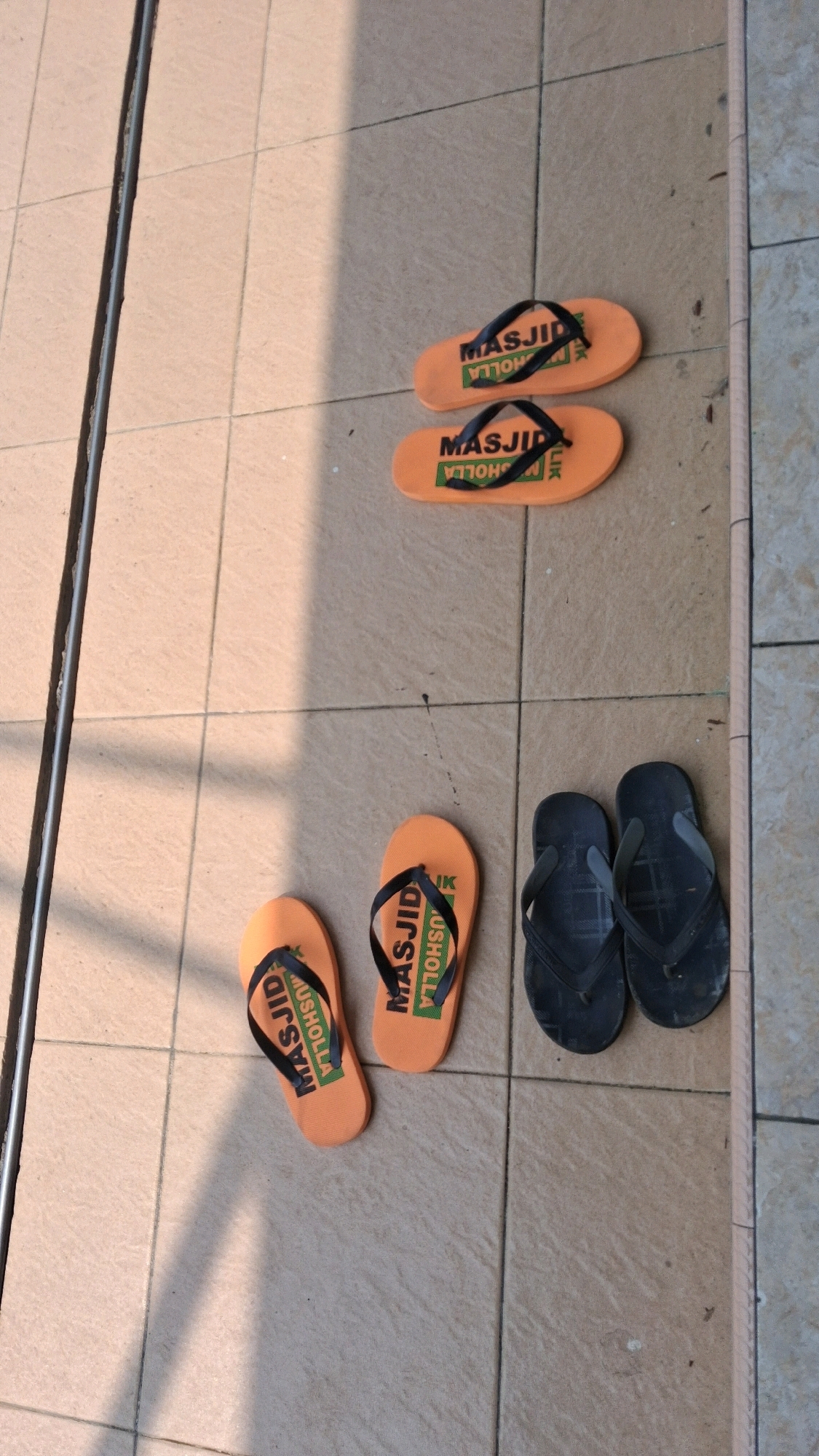 The image shows a top-down view of six pairs of flip-flops arranged on a tiled outdoor floor. The tiles are light brown with a subtle pattern. Sunlight casts a shadow across the left side of the image.
In the upper section of the image, two pairs of orange flip-flops are placed horizontally. Each has black straps and text printed on the soles. The text reads "MASJID" and "USHULLA".
In the lower section, there are two pairs of orange flip-flops placed diagonally, and one pair of black flip-flops placed horizontally. The orange flip-flops also have black straps and the text "MASJID" and "USHULLA" on their soles. The black flip-flops have grey straps and appear to be made of a rubber material.
The overall scene suggests a place of worship or a communal area where shoes are removed, possibly a mosque given the text on the orange flip-flops. The location is identified as Giriwarno, Indonesia.