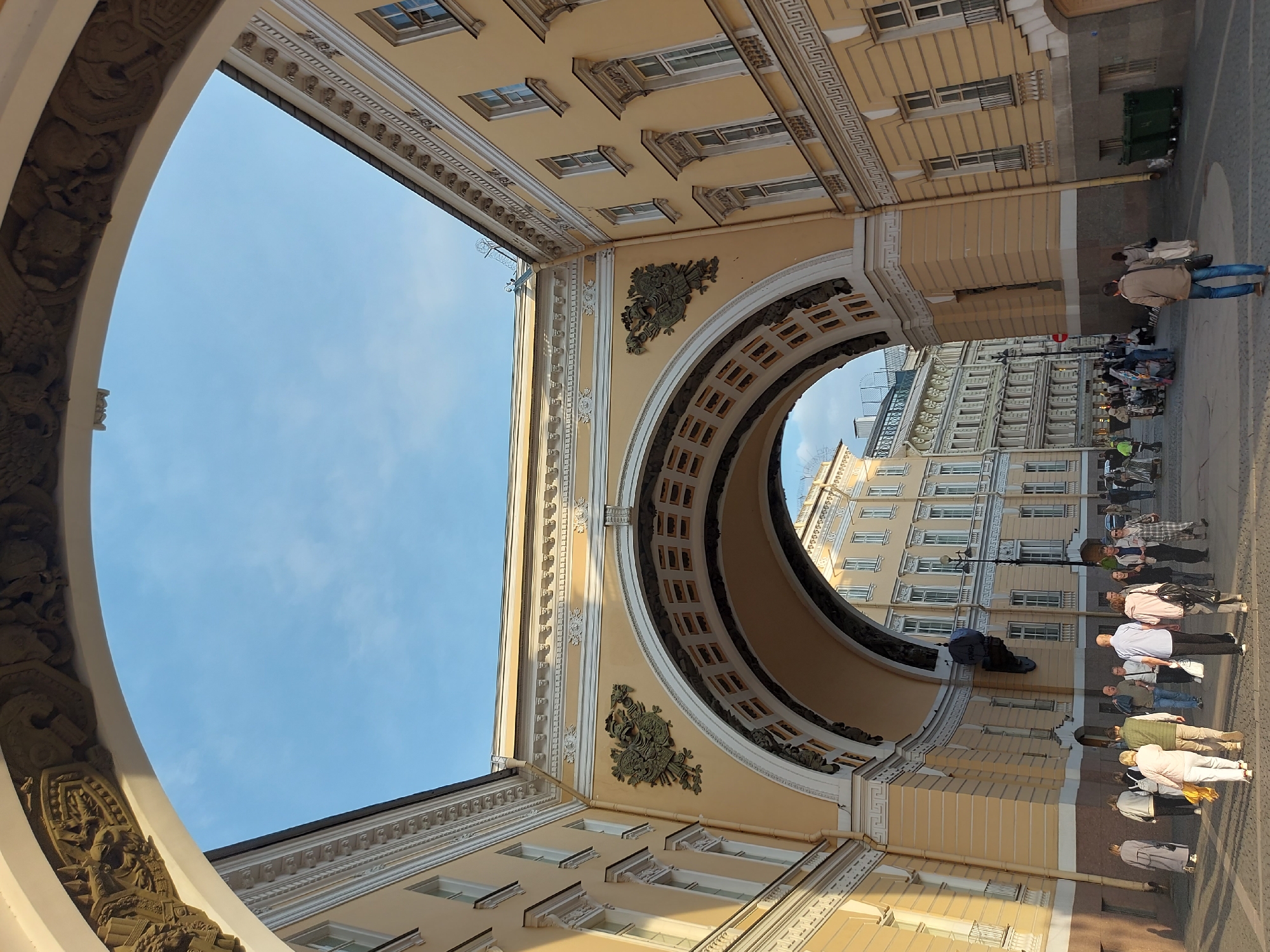 The image shows a view looking up through the arch of the General Staff Building in Saint Petersburg, Russia. The architecture is ornate, with decorative molding, columns, and classical details. The sky is visible through the archways, a clear blue. People are walking on the sidewalk outside the building. They appear to be dressed in casual clothing, some of them in summer attire. The foreground is dominated by the building's architecture, while the background features another building across the street, constructed in a similar architectural style. There are people walking across the street. There are also garbage receptacles on the sidewalk.