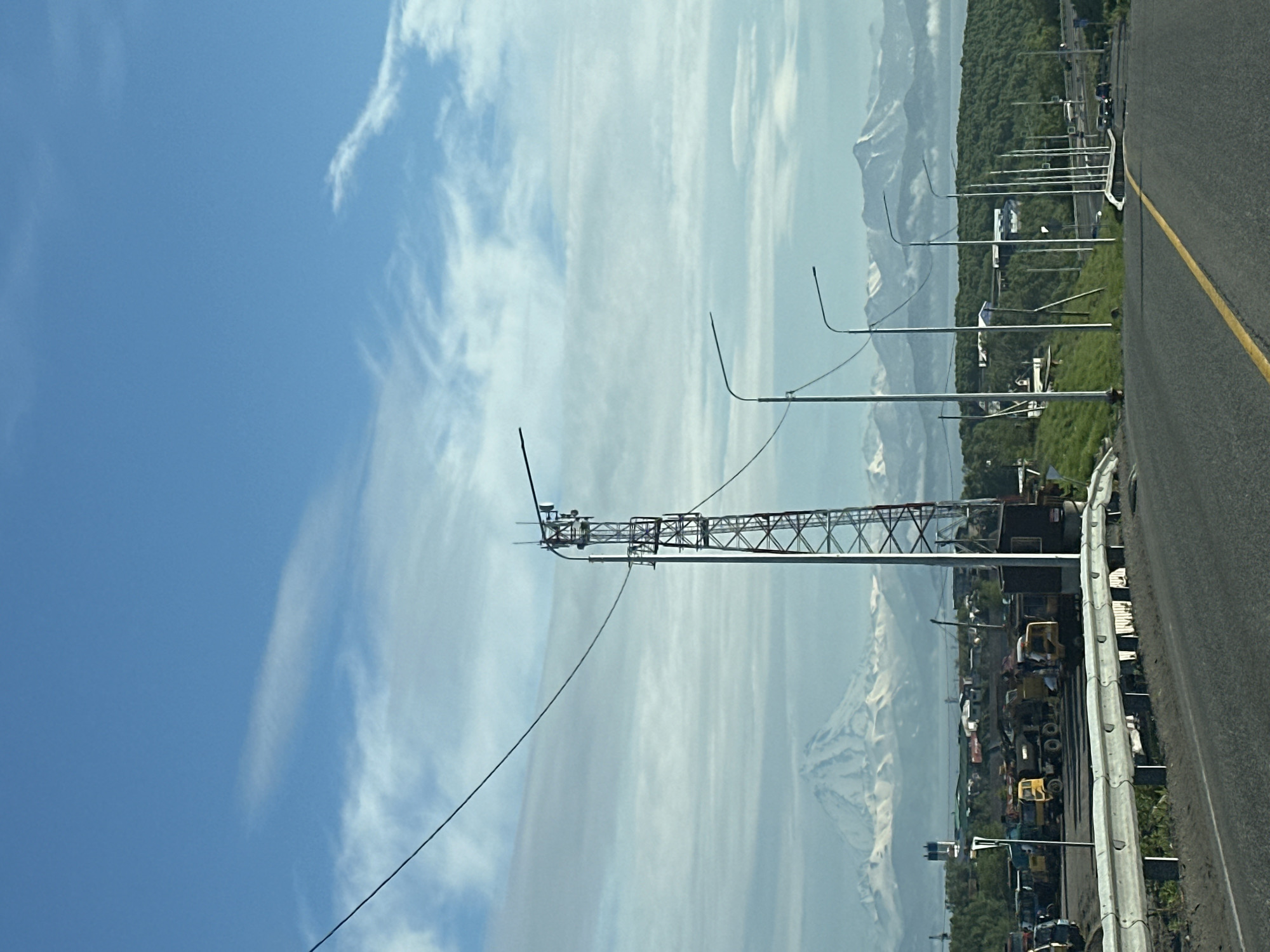 The image shows a view of a road leading towards a distant mountain range with snow-capped peaks under a partly cloudy blue sky. In the foreground, a paved road with a yellow dividing line curves to the right. A white guardrail runs along the left side of the road, with a grassy embankment behind it. To the left, a tall, lattice-style tower, likely for telecommunications, stands prominently. In the middle ground, there is a collection of heavy machinery, including yellow construction trucks and other vehicles, parked in an industrial or storage area. Beyond the machinery, several streetlights line the side of the road. Further in the distance, a treed hill rises, and beyond that, the snow-covered mountains are visible. The location is identified as Крутобереговый, Russia. There are no people or animals clearly visible in the image.