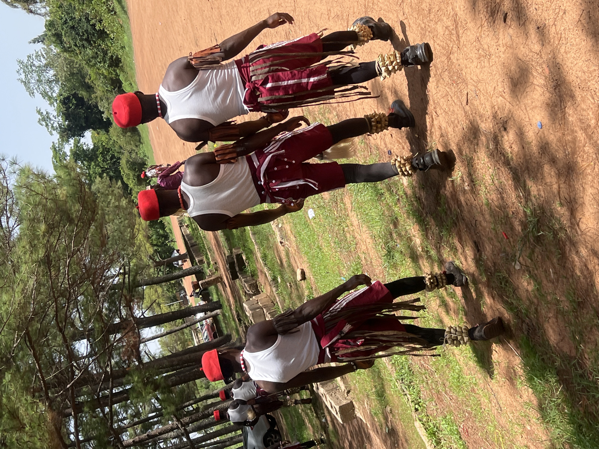 Three men wearing red caps, white tank tops, maroon shorts with white stripes, black leggings, arm protectors, and ankle ornaments are standing in a line. They are standing on grass and dirt under the shade of trees. The location is Jos, Nigeria.