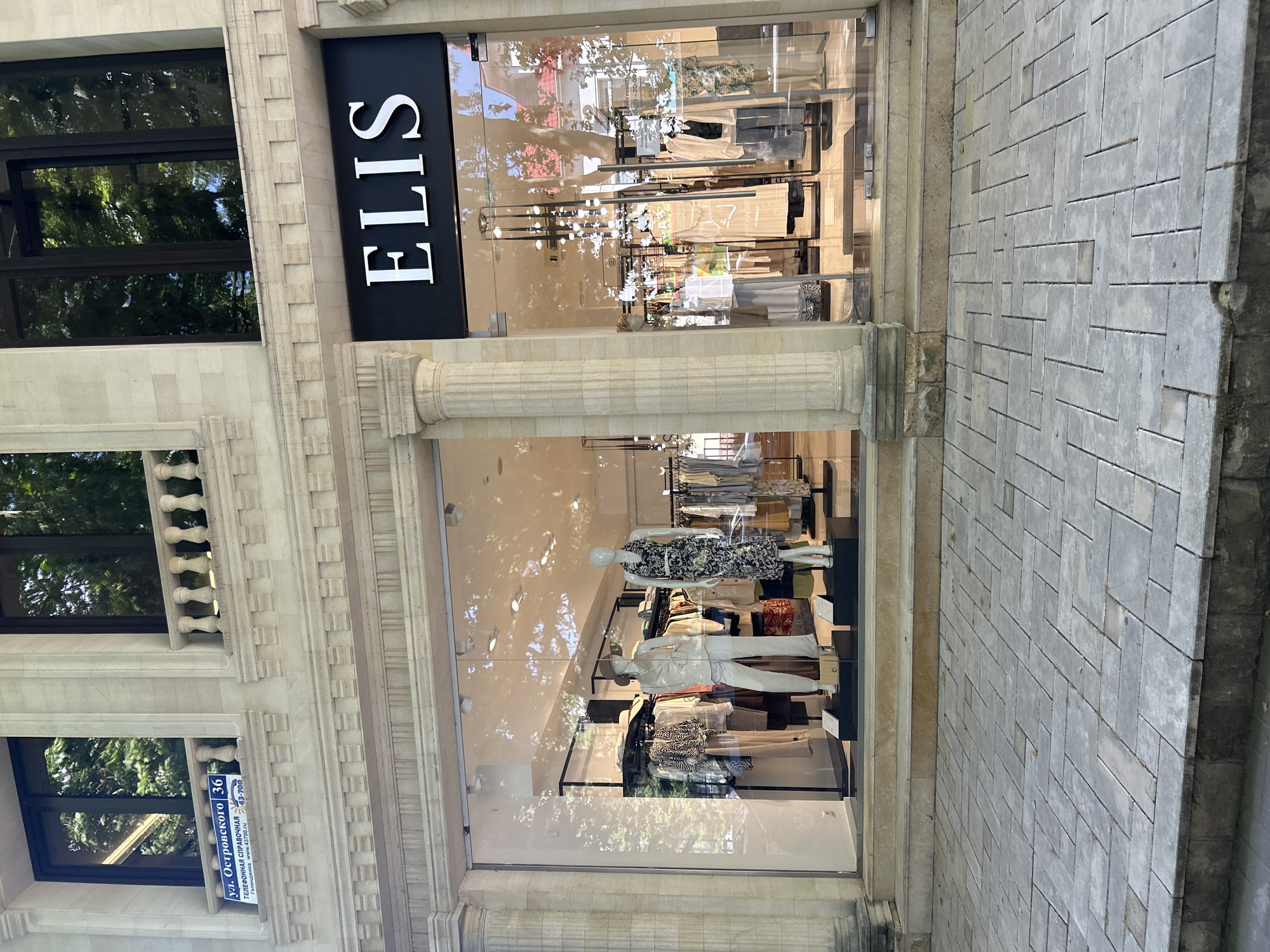 The image shows the exterior of a store called "ELIS" with a large glass window displaying clothing and mannequins. The store is located on a street with a cobblestone sidewalk. The building is made of stone with architectural details like columns and decorative moldings. Sunlight filters through trees, creating dappled shadows on the facade and sidewalk, suggesting it's daytime. The store window features a fashion display with several mannequins dressed in various outfits, including dresses, pants, and tops. Racks of clothing are also visible inside the store, indicating a retail environment. A blue sign with Russian text is attached to the building near a window. The text on the sign reads "ул. Островского 36", "Телефонная справочная 43-700", and "www.43700.ru". The overall scene depicts a typical urban streetscape with a focus on a fashion boutique.