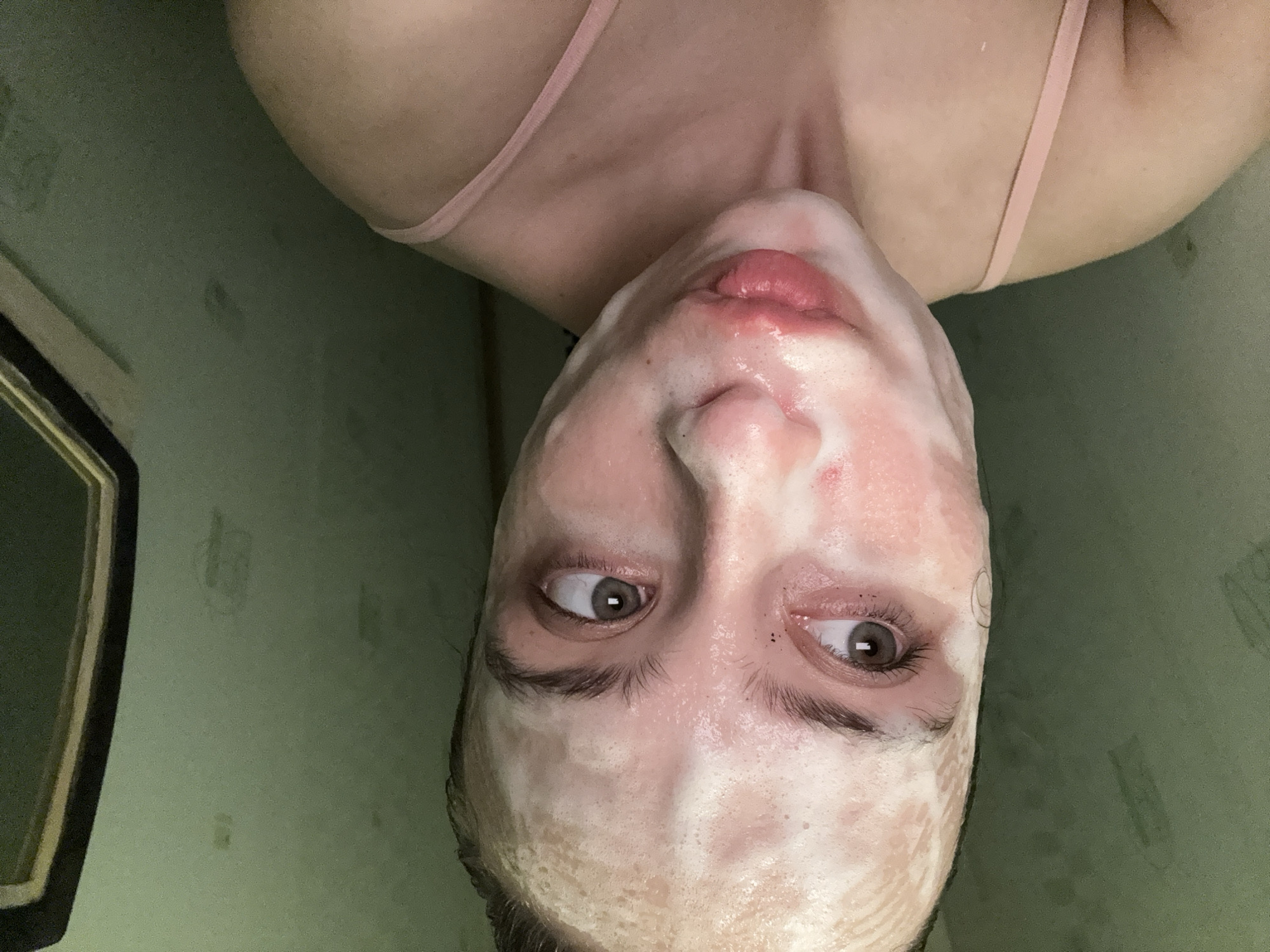 A woman in Прикамский, Russia with a facial mask is photographed close-up. The background is a green wall with marks. A mirror is slightly visible on the left. The woman has fair skin, brown eyes, and brown eyebrows. She wears a pink tank top. Her face is covered in a white mask, and her lips are slightly parted.
