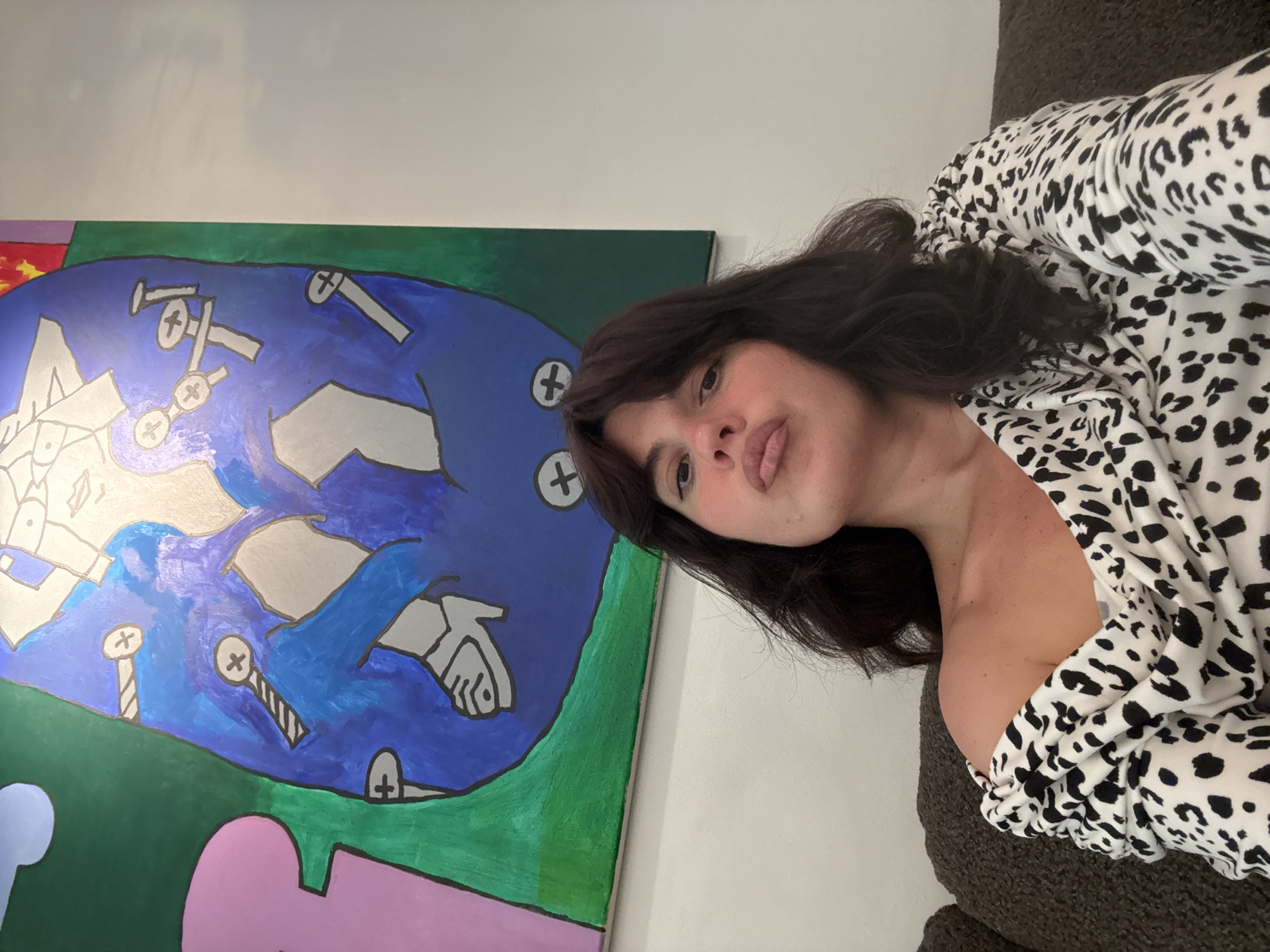 A woman with dark hair and fair skin is lying on a dark gray upholstered surface, wearing a white and black spotted blouse. A colorful painting hangs on the wall behind her, featuring a predominantly blue background with abstract shapes and images resembling screws or nails. The painting also incorporates green, pink, and red sections. The scene takes place indoors, with a white wall visible in the background. The location is likely a private residence, but no specific details indicate that it is located in Berlin, Germany.