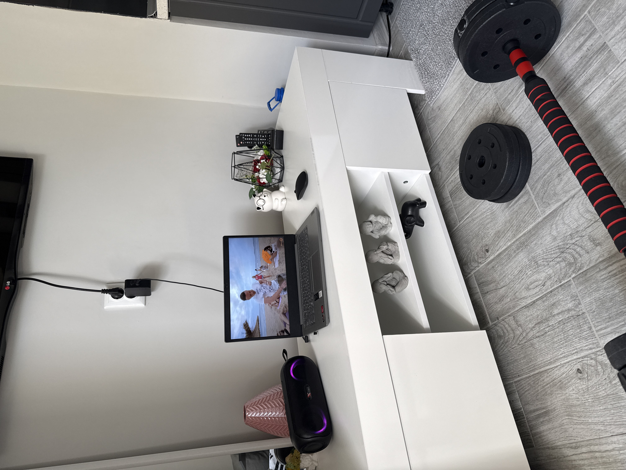 The image shows a room with a television mounted on the wall, plugged into a wall outlet. Adjacent to the TV is a white storage unit with open shelves. On the surface of the unit, there is a laptop, a speaker, decorative items like a pig figurine and a floral arrangement, and a remote control. Inside the shelves are multiple gray sculptures. Beside the storage unit, a barbell with weights is leaning against the wall. The flooring is gray wood-look tiles. Based on the objects in the image, it is difficult to provide specific details about the location in Stara Zagora, Bulgaria. There are no people visible in the image.