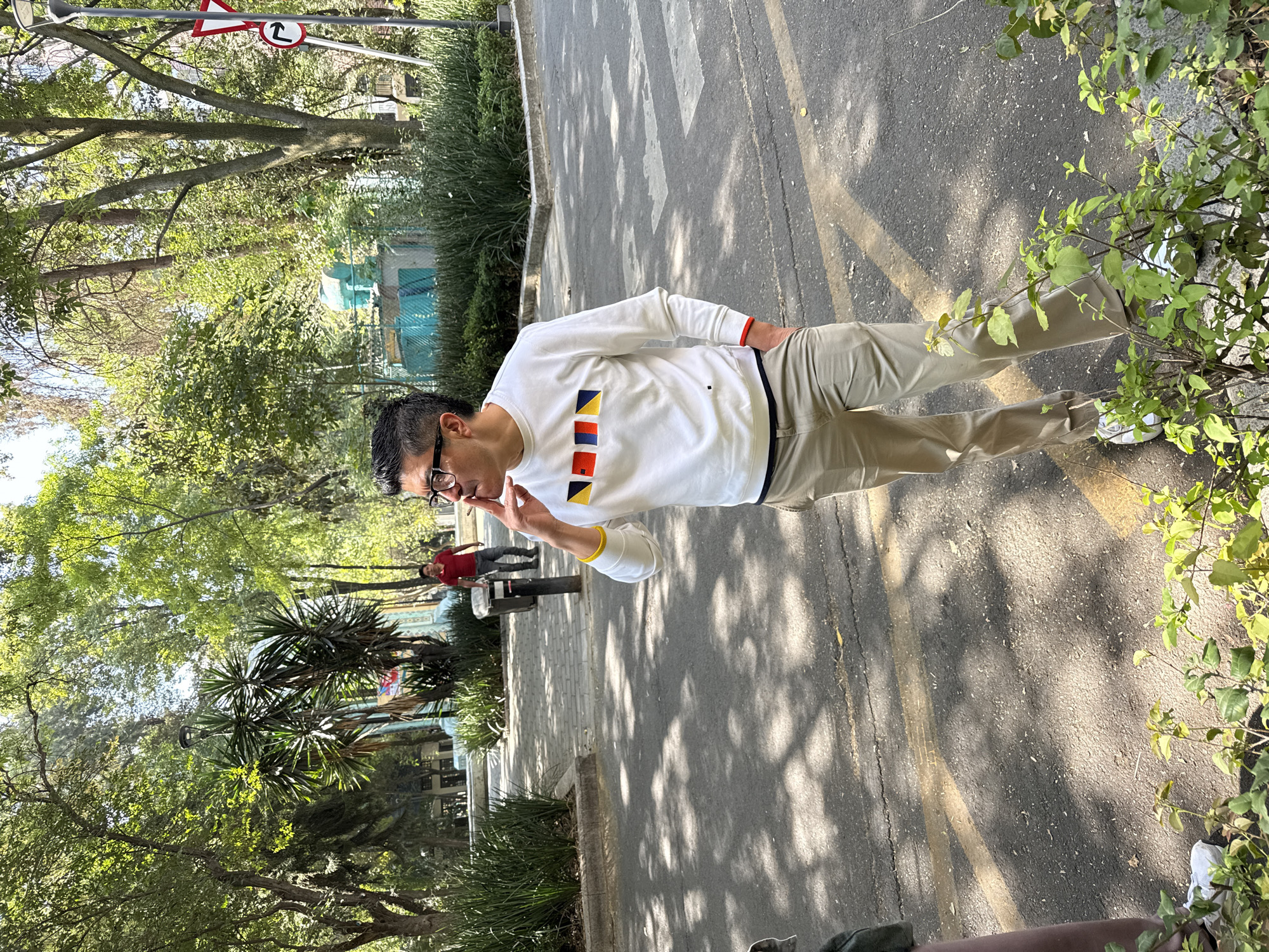 A man in glasses and light-colored clothing stands on a gray asphalt surface, seemingly a sidewalk or street. The scene is heavily shaded by trees, casting dappled light across the ground. He is holding a cigarette in his hand. In the background, another individual in a red shirt is visible. Vegetation and trees are abundant, lining the edges of the walkway. A street sign is present in the upper left corner. The location is Mexico City, Mexico.