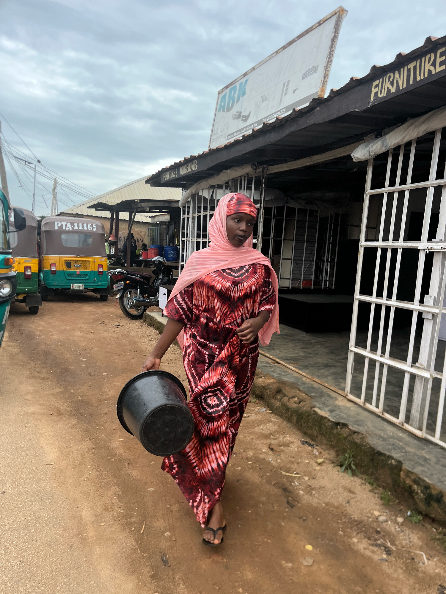 In Jos, Nigeria, a young woman walks down a dirt road carrying a black bucket. She is wearing a pink headscarf and a long, patterned dress. To her right, a store with a sign that reads "FURNITURE" is visible, with a white metal gate. In the background, several auto-rickshaws are parked on the side of the road. The sky is overcast.