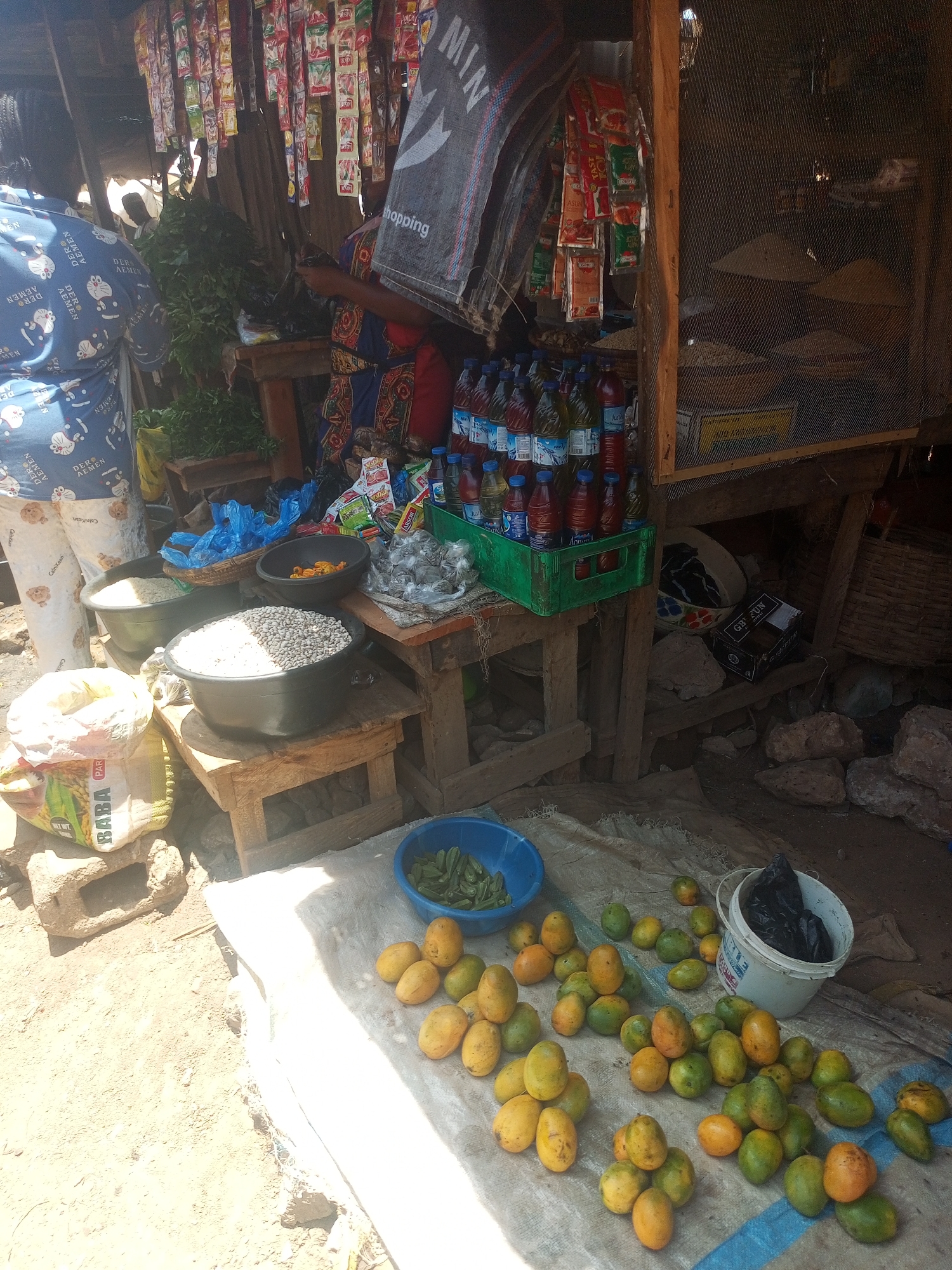 A roadside market stall in Sabon Tasha, Nigeria, displays various goods. A person wearing a blue patterned shirt stands at the left, while another in a blue and red patterned dress stands behind a wooden counter stocked with items like seasoning packets, bottled drinks in a green crate, and bags of rice, beans and other dry ingredients in black bowls. On the ground, mangos and okra fill a sheet and blue bucket. A white bucket sits behind them.