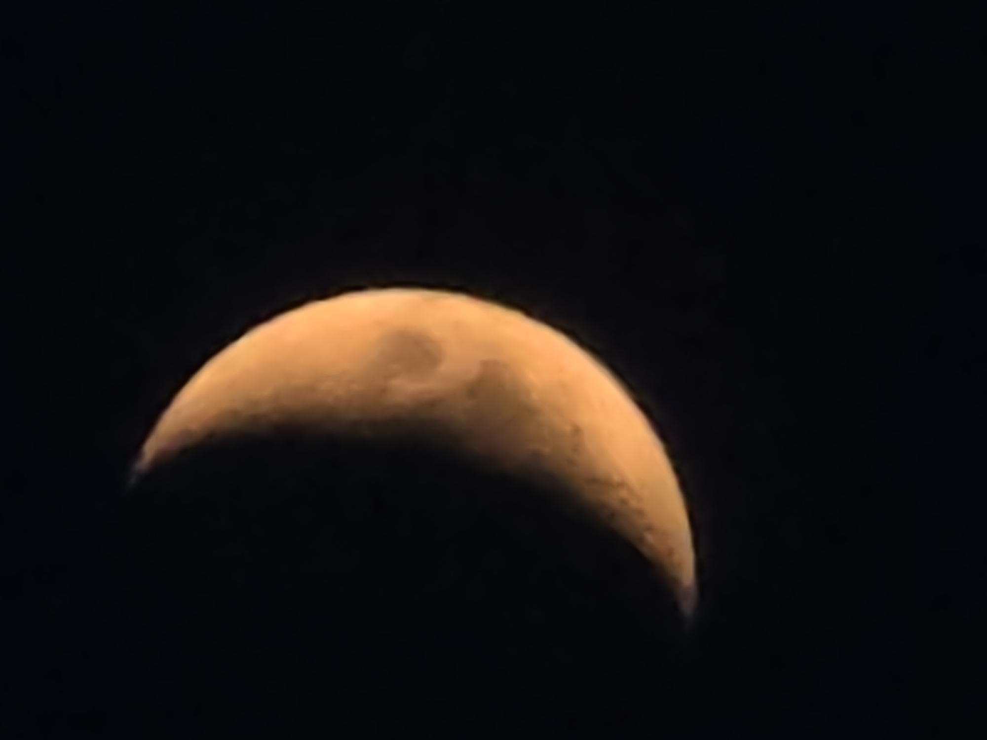 The image shows a crescent moon against a black sky. The moon is illuminated from the right, with the illuminated portion being a bright orange color. The unilluminated portion is dark and blends into the background. There are some subtle variations in shading on the illuminated part of the moon, suggesting craters or other surface features. The image is tightly cropped on the moon, with no other objects or features visible. There is no information provided about the location of this image in Podgorica, Montenegro.
