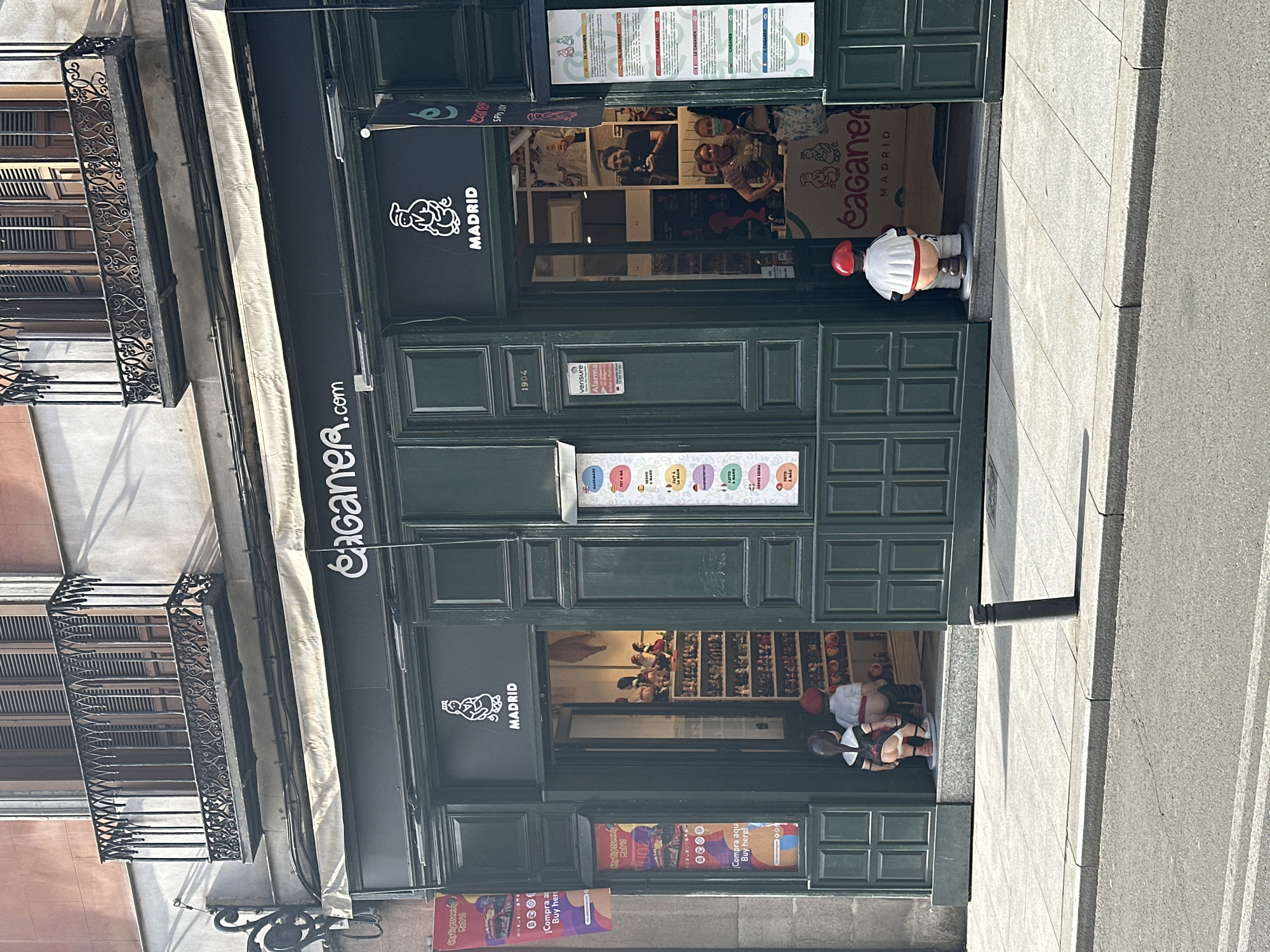 Here is a detailed description of the image:
**Scene Overview:**
The image shows the storefront of a business named "Caganer.com" located in Madrid, Spain. The building appears to be an older structure with a multi-story façade.
**Storefront Details:**
* **Signage:** Above the door, there's a sign with "Caganer.com" and a logo featuring a squatting figure (a caganer). Additional signs advertising "Caganer Madrid" are also visible.
* **Exterior Decor:** To the right of the door is a statue of a caganer wearing a chef's hat.
* **Doors:** The storefront has green doors with divided panels. There are promotional posters displayed in the lower sections of the doors.
**Interiors:**
* Through the upper door, customers and staff can be seen. Boxes with "Caganer Madrid" are visible in the background.
* Through the lower door, shelves display figurines, indicating the type of product sold in the store.
**Contextual Elements:**
* **Architecture:** The building has wrought-iron balconies on the upper floors.
* **Street:** A section of sidewalk is visible, suggesting the store is located on a busy street.
* **Signage:** there are City Sightseeing Madrid posters on the store front.
**In summary:** The image showcases a shop specializing in caganers, a traditional Catalan Christmas figurine, in Madrid.
