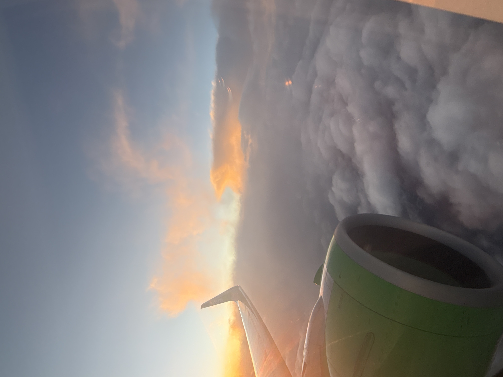 The image depicts a view from an airplane window, showing the wing and engine of the aircraft in the foreground. The wing is silvery and partially illuminated by sunlight. The engine casing is green and has visible details like bolts and seams. Beyond the aircraft components, the sky is visible. Part of the sky is a clear blue, transitioning into clouds. Some clouds have a golden hue, likely due to the setting or rising sun. Other cloud formations are dense and gray, resembling a storm. The image is taken during flight, with the emphasis on the natural beauty of the sky and clouds viewed from above. The location is unknown, but it is not likely Mexico City because the weather conditions shown are not typical for this city.