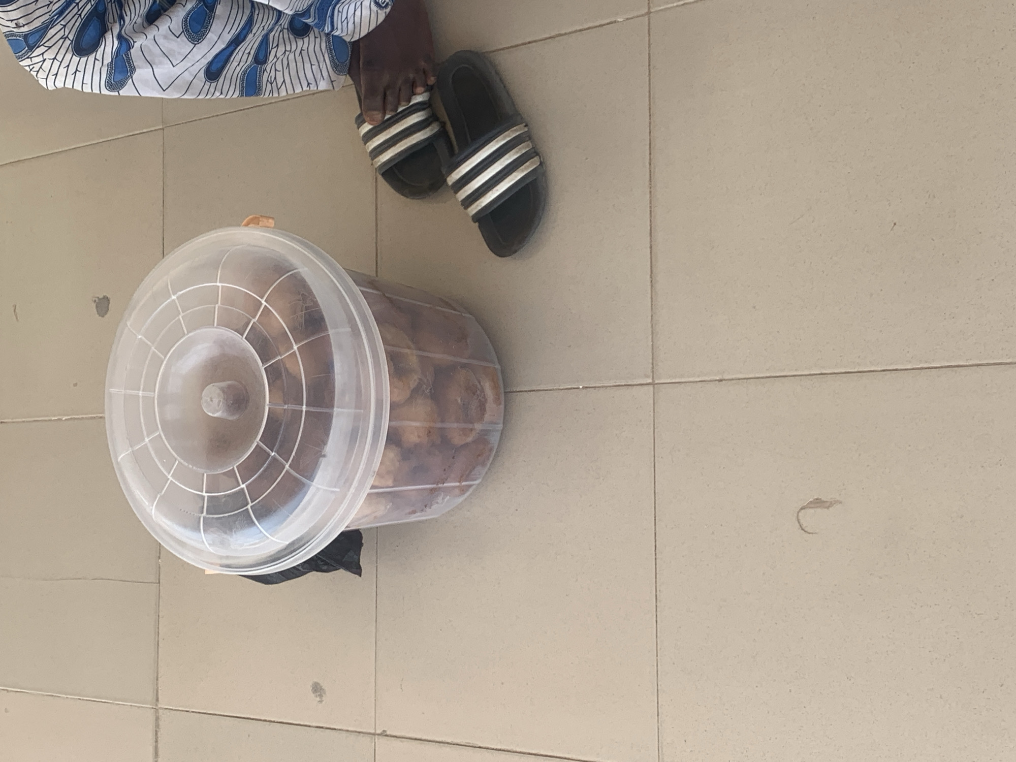 The image shows a close-up, overhead view of a person's feet and a clear plastic container filled with food items, resting on a tiled surface. The person is wearing black slide sandals with three white stripes. The person's left foot is visible, with toes peeking out from under the sandal. They are wearing a white and blue patterned fabric around their lower legs.
The plastic container is large and cylindrical, with a domed, transparent lid that has a concentric ring pattern. Inside the container, numerous round, brown food items are visible, appearing to be a snack or pastry. The container is placed on light-colored floor tiles, which are arranged in a grid pattern. A small piece of black material, possibly a bag, is partially visible underneath the container.
The setting appears to be outdoors or in a covered outdoor area, given the lighting and the tiled floor. The time of day is not explicitly indicated, but the lighting suggests it could be daytime. There are no visible signs of weather conditions. The image provides no explicit text. The focus is on the everyday elements of a person preparing to share or store food. The overall impression is a candid, unposed moment.