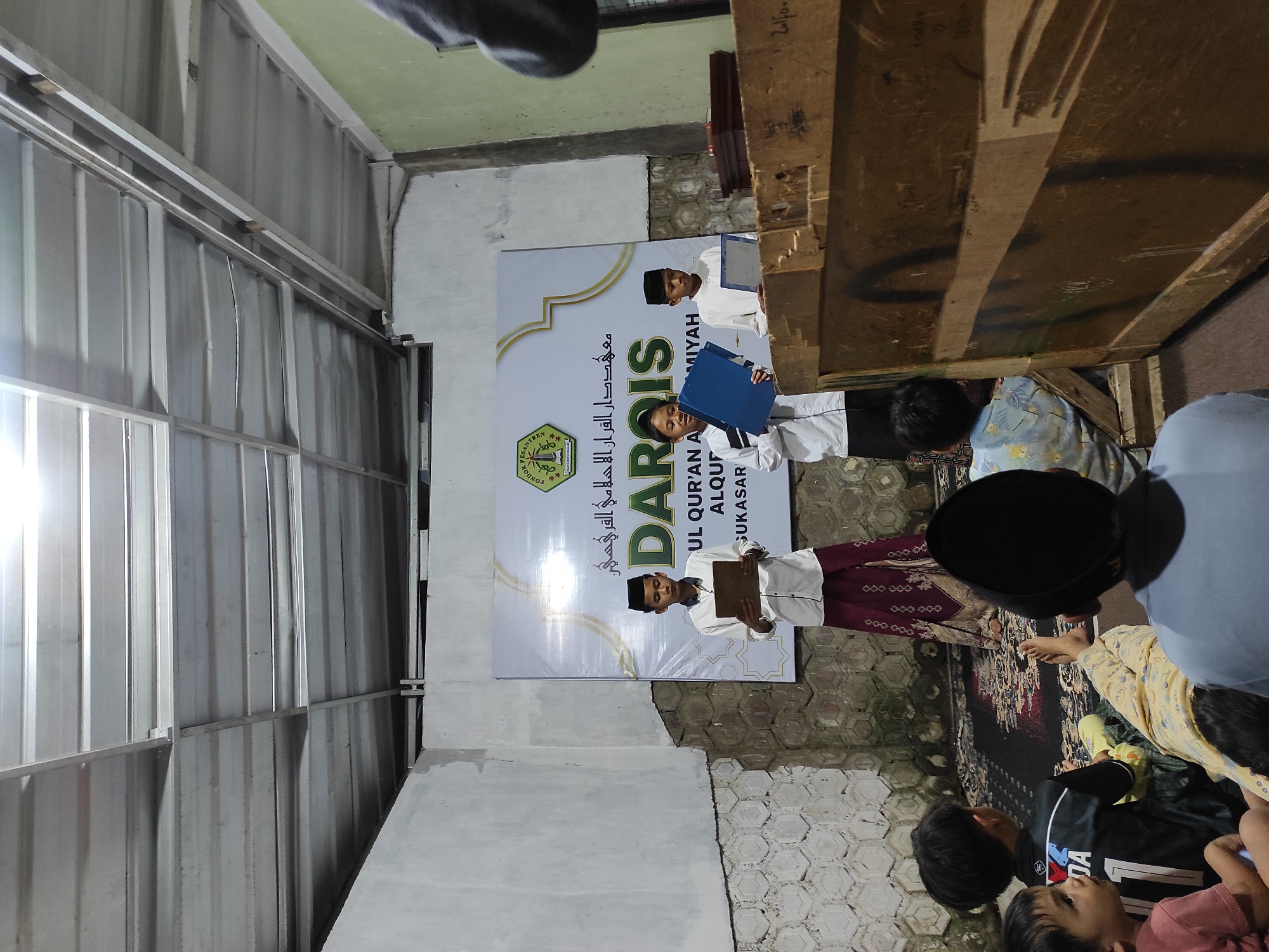 The image depicts an indoor event with several children and teenagers. In the background, a banner with Arabic script and the text "PONDOK PESANTREN UL QUR'AN AL-QUR'AN AL-MIYAH SUKASARI" is visible, indicating the event is likely taking place at a pesantren (Islamic boarding school) in Sukasari, possibly in the Sumedang region of Indonesia, given the Arabic script and context.
Three young male students stand on a stage-like area, facing forward. The student on the left is holding a wooden tablet. The central student is holding a blue folder. The student on the right is holding a tablet or book. They appear to be participating in a recitation or presentation.
In the foreground, several younger children are seated on the floor, facing the stage. They are dressed in casual attire and appear to be an audience. One child wears a black jersey with the number "1" and the word "DA". Another child wears a blue t-shirt.
The environment is simple, with a corrugated metal ceiling and concrete walls. A large wooden structure, possibly a crate or makeshift stage, is positioned to the right of the frame. The lighting appears to be artificial, casting shadows. The floor is covered with a patterned rug. The overall scene suggests a modest community or educational event.