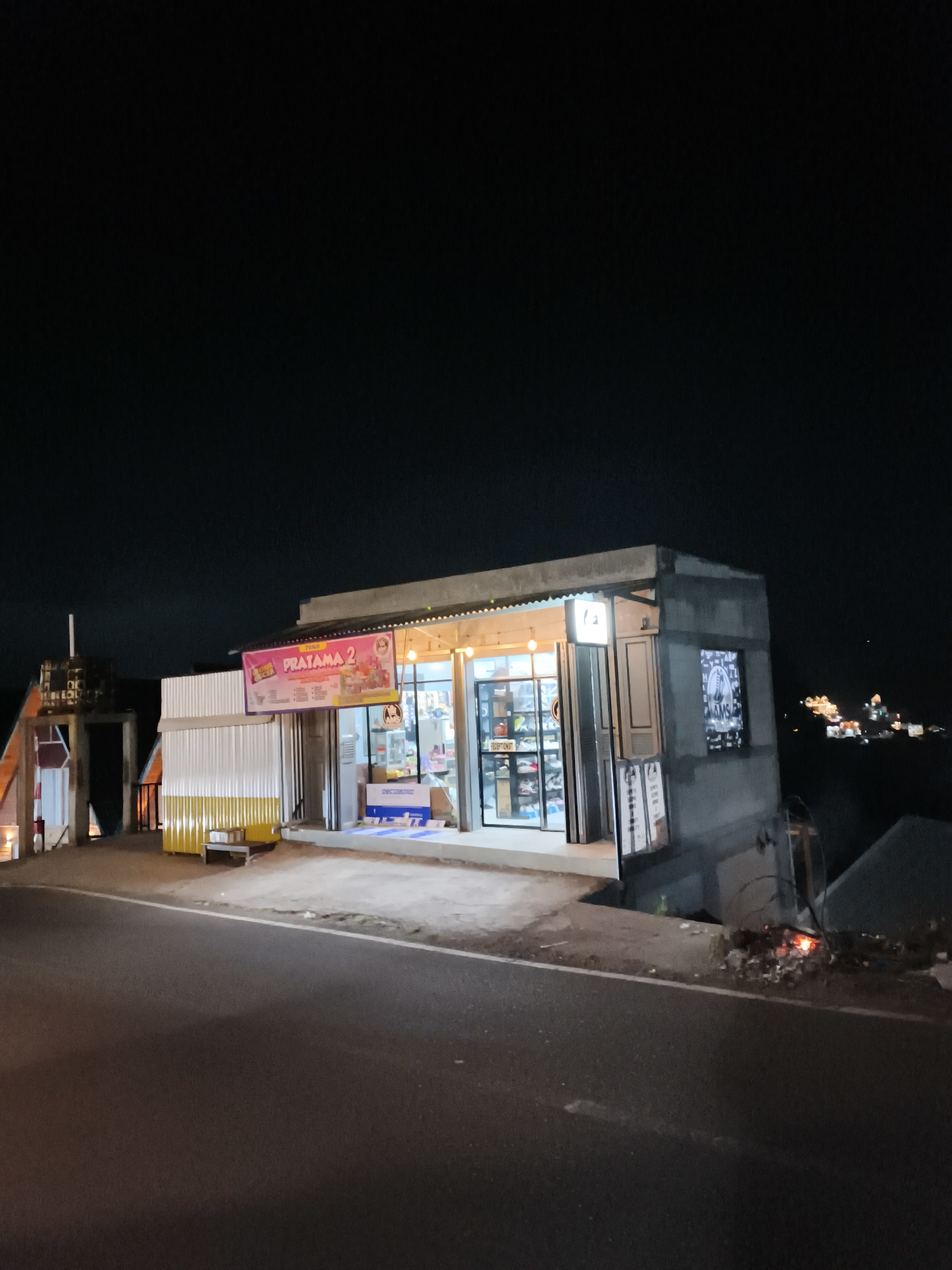 The image shows a small convenience store, likely a "warung," at night in Dieng Kulon, Indonesia. The store, named "PRATAMA 2," is illuminated by interior lighting, showcasing shelves filled with products visible through glass doors. A banner with colorful graphics and text is displayed above the entrance. To the left of the store, there's a structure with a water tank on top and a yellow and white corrugated metal enclosure with some boxes on a bench. A road is in the foreground, with a dark, unlit landscape and distant lights of a town or settlement visible on a hillside in the background. The scene appears to be on the outskirts of the main settlement, given the darkness and the exposed road. There are no people visible in the image.