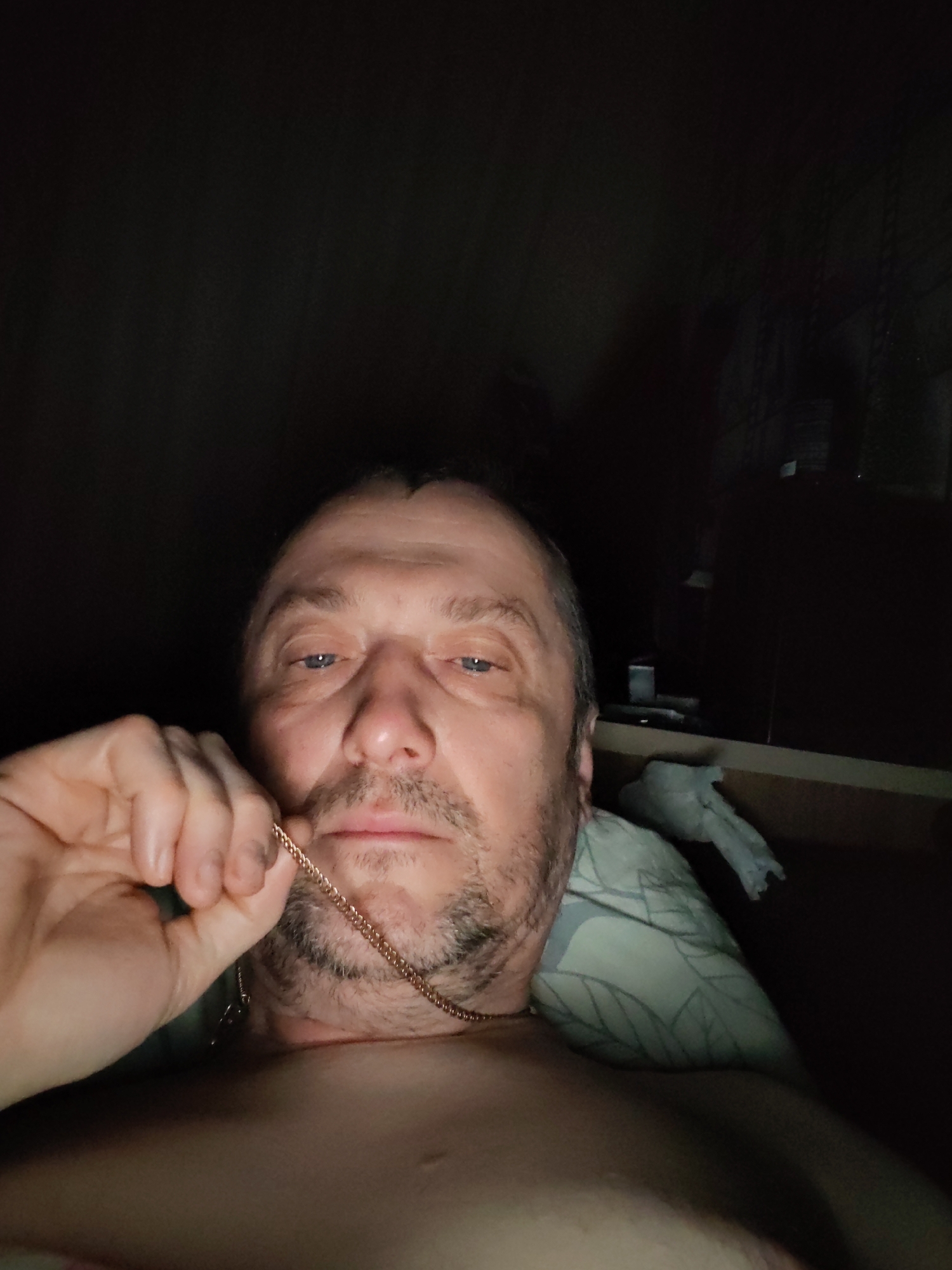 A Caucasian male with a light complexion, stubble, and a receding hairline is positioned in the foreground, holding a gold chain near his face. He appears to be lying down, possibly in a bed, as suggested by the pillow visible behind him. The background is dark, with only a faint outline of what appears to be a window or a shelf with items on it. The overall lighting is dim. The location in the city of Chaikovsky, Russia, is unknown.