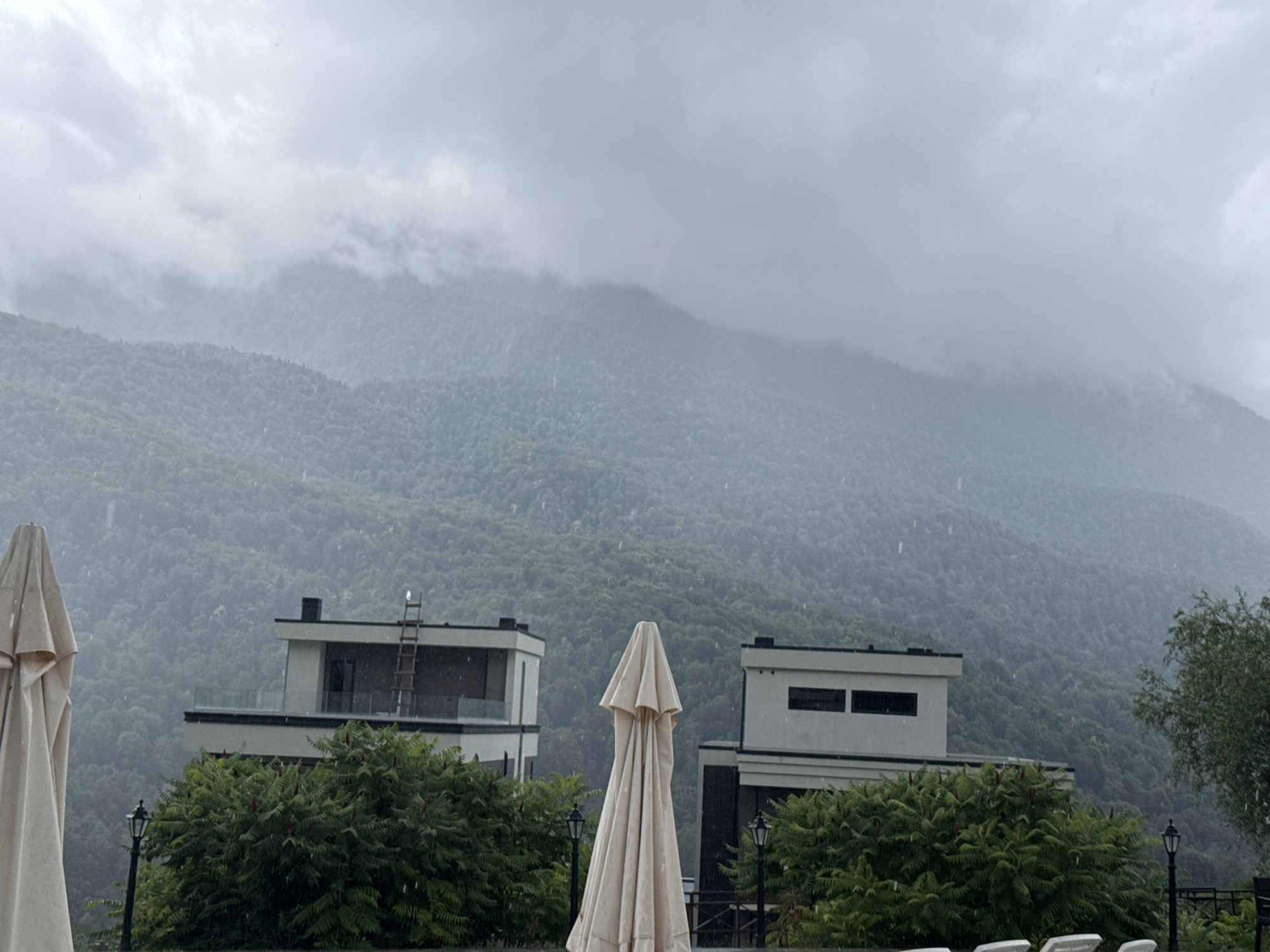 The image depicts a moody, overcast day in Krasnaya Polyana, Russia, with rain falling against a backdrop of lush, forested mountains. In the foreground, two modern buildings with minimalist architecture stand amidst dense green foliage. To the left, a large, cream-colored umbrella is folded and tilted, partially obscuring the view of one of the buildings. Another identical umbrella is positioned in the center of the frame, also closed and upright, creating a somewhat eerie, ghost-like appearance. Streetlights are visible among the bushes, and the rain can be seen as faint streaks across the image. The mountains in the distance are shrouded in mist and clouds, adding to the somber atmosphere. The scene suggests a tranquil, yet slightly melancholic, mountain resort setting during inclement weather.