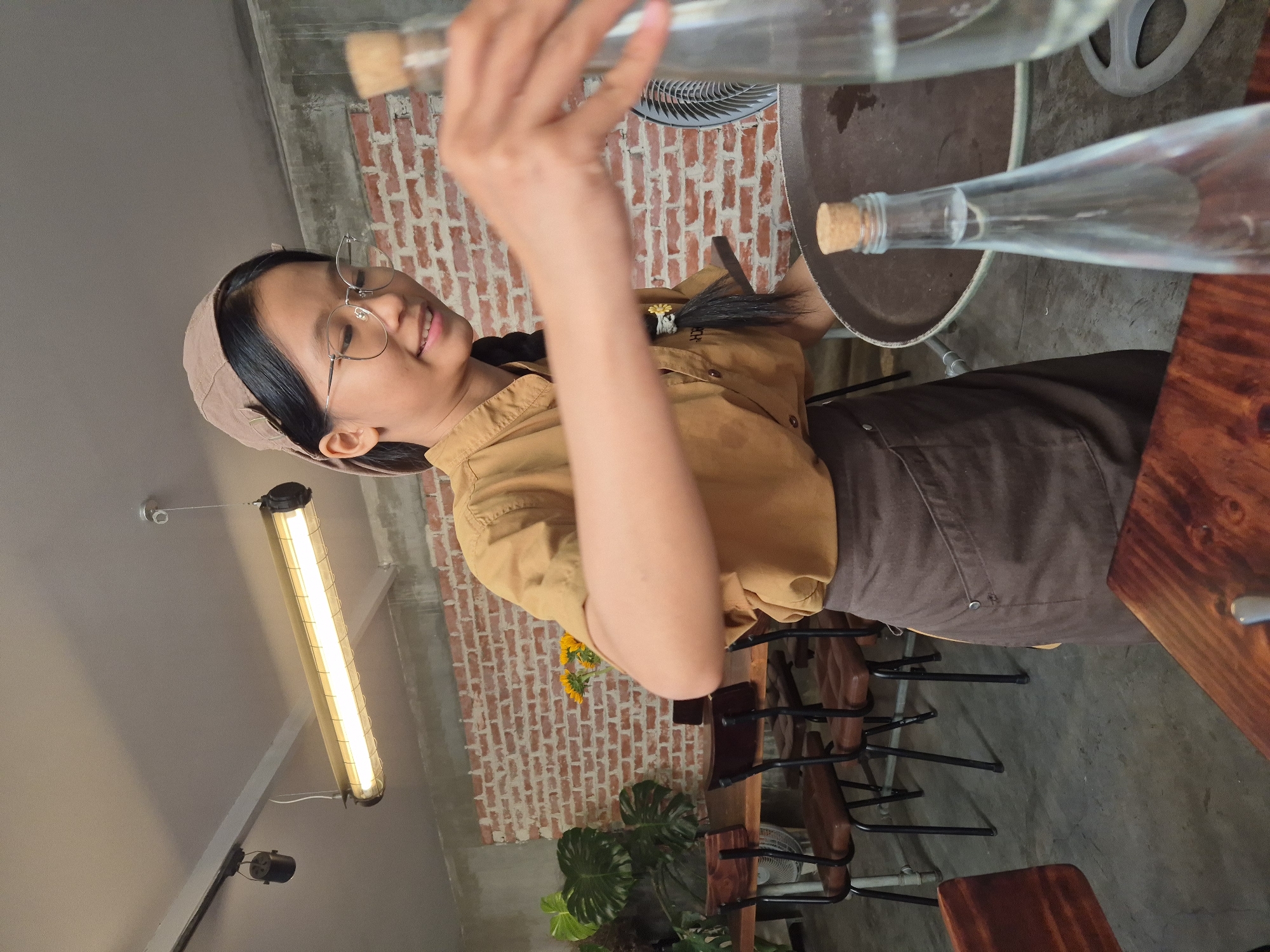 A young Vietnamese woman wearing a tan cap, glasses, and a tan uniform with a brown apron is serving drinks at a restaurant. She has a black braid with a yellow flower accessory. She is holding a round tray with two glass bottles of water. The restaurant has exposed brick walls, a long fluorescent light fixture on the ceiling, wooden tables and chairs, and potted plants. The image appears to have been taken in Da Nang, Vietnam.