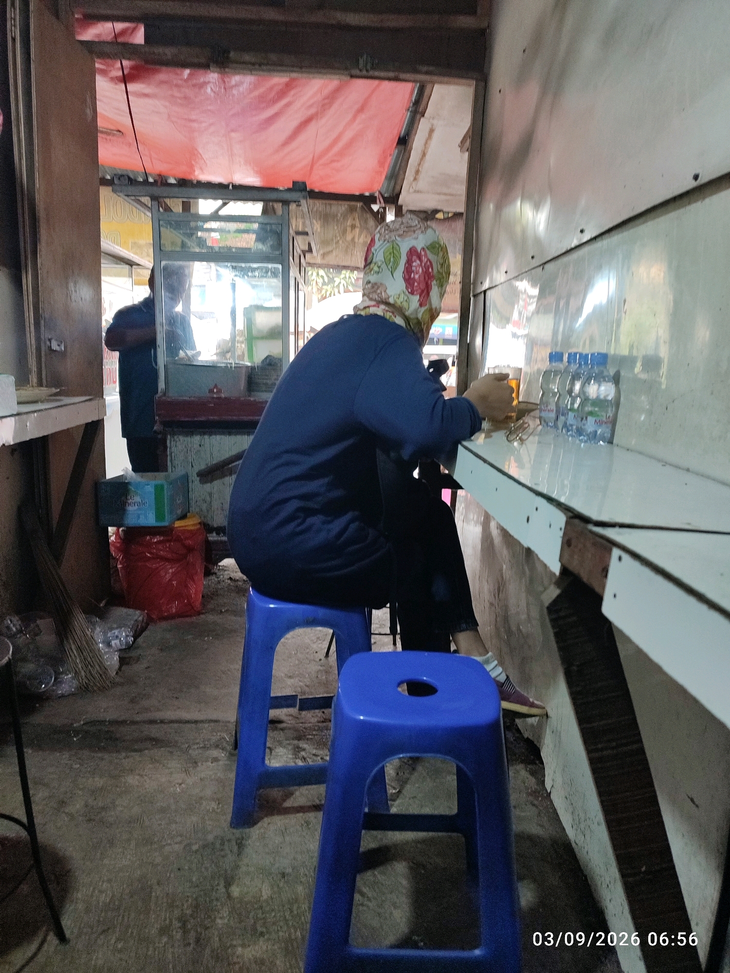 A person wearing a blue long-sleeved shirt and a patterned headscarf is seated on a blue plastic stool at a white table in what appears to be an open-air food stall or small eatery. They are holding a glass of amber liquid and are facing away from the camera, towards a counter area. On the table, there are several plastic bottles of water and a pair of eyeglasses. Behind the person, a second individual is visible in the reflection of a glass partition, wearing a dark blue garment. The environment is rustic, with visible structural elements of the stall, including a red tarpaulin overhead. On the floor, there is a red plastic bag, a blue cardboard box, and a broom. The scene is captured with a timestamp of "03/09/2026 06:56". Based on the visual cues, this scene is likely located in East Jakarta, Indonesia, characteristic of a local warung or kedai kopi.
