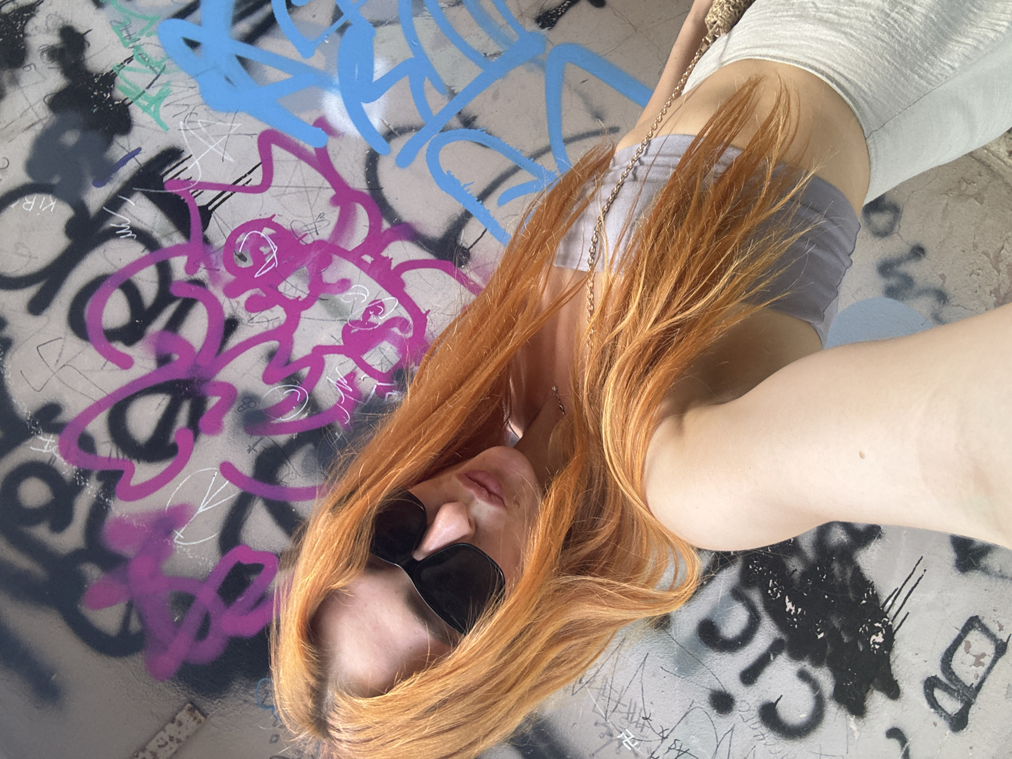 A woman with bright orange hair, wearing sunglasses, a grey tube top, and a light-colored skirt, poses for a selfie against a graffiti-covered wall. The graffiti features various colors, including pink, blue, black, and white, creating a vibrant and urban backdrop. The woman's pose suggests a casual and confident demeanor. The image captures a moment in what appears to be an outdoor urban environment, likely in Budva, Montenegro, given the context of the city.