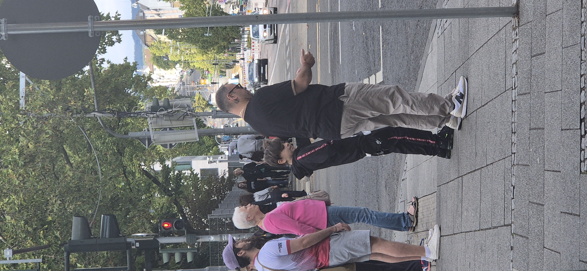The image captures a street scene in Wiesbaden, Germany. In the foreground, four individuals are visible on a paved sidewalk. From left to right, a man wearing a purple cap, a white t-shirt with red trim, and grey shorts stands facing left. Next to him, a woman with short white hair, wearing a bright pink long-sleeved shirt and blue jeans, faces left, with a light-colored shoulder bag. A young boy, dressed in a black hooded sweatshirt with red stripes on the sleeves and black pants with red stripes down the side, stands facing right, looking at the man beside him. The man to the boy's right, wearing glasses, a dark t-shirt, and light beige pants, is also facing right and appears to be holding something in his right hand, possibly an object or his fingers.
Behind this group, several other people are visible, some walking further down the sidewalk. The environment includes a traffic light displaying a red signal, trees with green foliage, and buildings in the background. The street features cars and what appear to be road construction barriers. The perspective suggests the photo was taken from across the street or at an intersection.